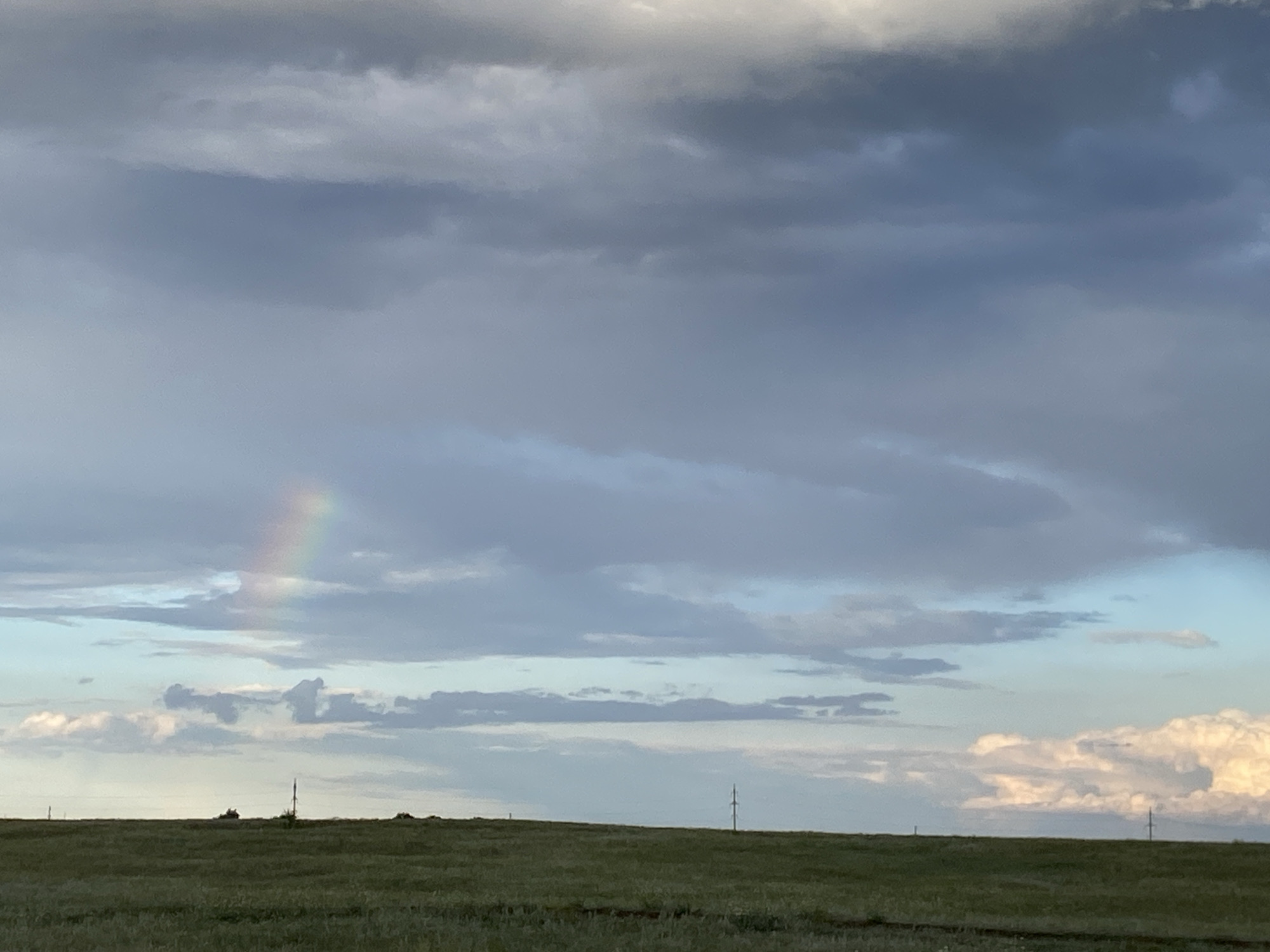 A wide shot captures a grassy, undulating field under a vast sky. A faint rainbow arcs across the upper left portion of the frame, set against a backdrop of layered clouds ranging from dark gray to lighter hues with hints of blue and white near the horizon. A few sparse, dark, vertical structures, possibly utility poles or small trees, dot the distant treeline. The ground is covered in green grass, suggesting an outdoor, rural or undeveloped setting. The lighting indicates either early morning or late evening, with a soft, diffused light illuminating the scene. The location is identified as Байқоныс ауылдық округі, Kazakhstan.