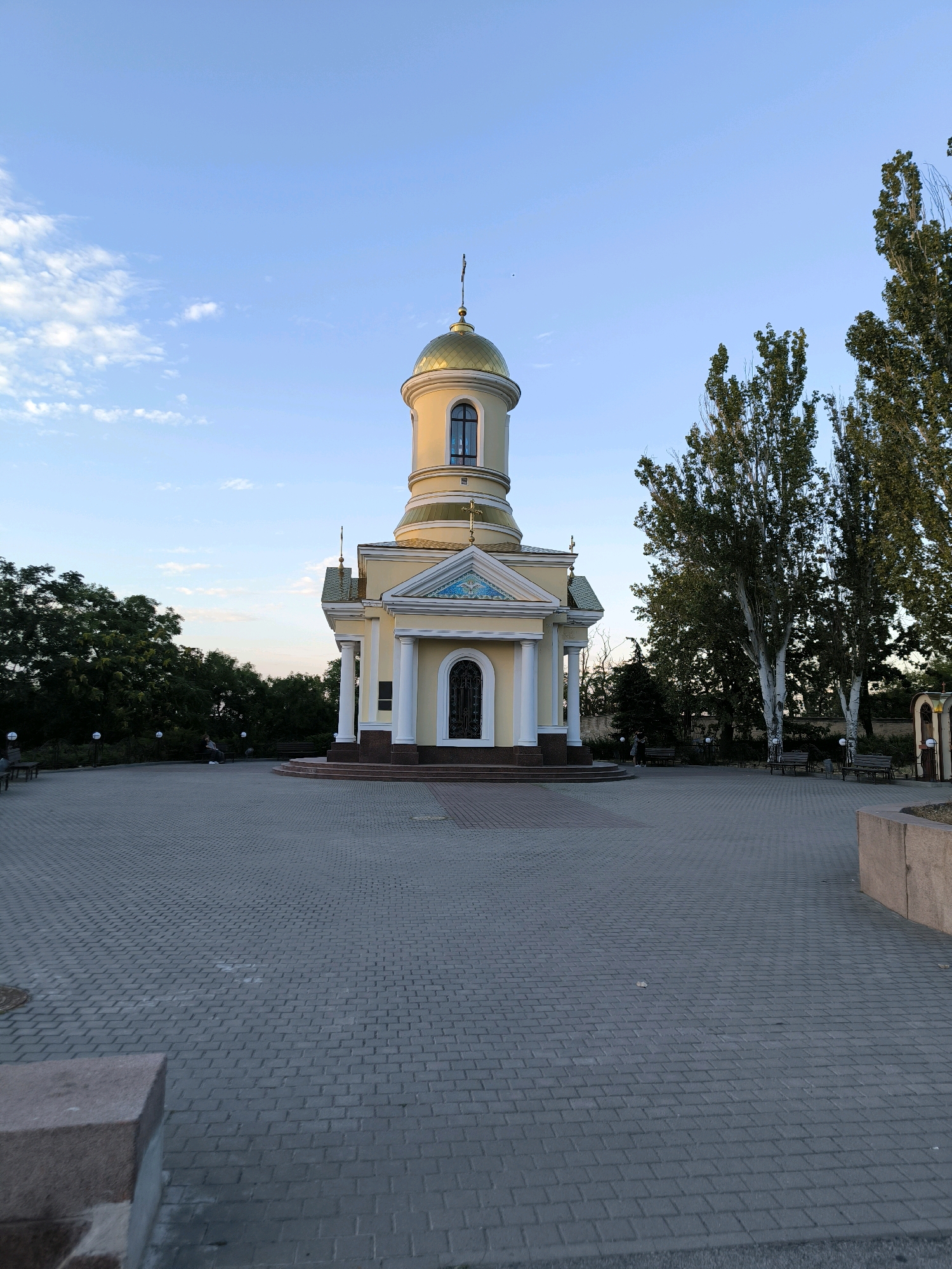 The image depicts a small, yellow chapel with a golden dome, set against a clear blue sky with sparse clouds. The chapel features white columns, a decorative frieze above the entrance, and an arched window on the dome. The foreground is a paved plaza with a brick pattern. To the right of the chapel, tall, slender trees with light-colored bark stand out against a backdrop of darker foliage. Benches are visible in the plaza, and a few people are seated on them. In the distance, more trees and a fence line can be seen. The scene appears to be captured during daylight, possibly late afternoon given the lighting. This is the chapel of St. Nicholas the Wonderworker, located in the city of Mykolaiv, Ukraine, specifically at the memorial complex dedicated to the naval victory of 1788.