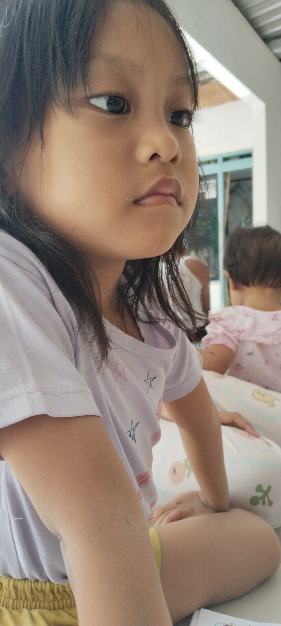 The media file is a medium close-up shot of a young girl, captured from the chest up, in a horizontal frame. She is looking to her left with her mouth slightly ajar. Her dark hair frames her face, and she is wearing a light purple t-shirt with a pattern of stars. In the background, another child, likely younger, is visible wearing a pink outfit with ruffles. The setting appears to be indoors, possibly a home, with hints of windows and ceiling in the background. The lighting suggests it is daytime. The focus is primarily on the girl in the foreground, highlighting her facial features and attire.