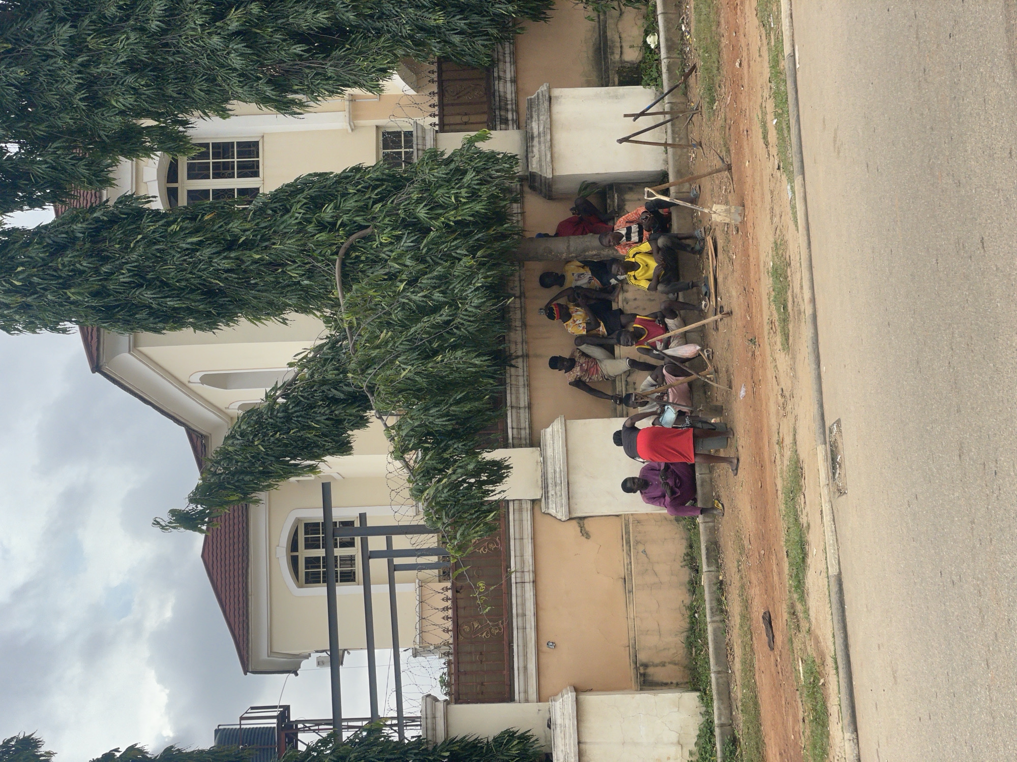 In Guzape, Nigeria, a group of men are gathered under a tree on a dirt patch next to a paved road. The men are casually dressed, with some wearing t-shirts and shorts, and others in traditional attire. They are seated on a low wall and on the ground, some appearing to be resting or engaged in conversation. Several gardening tools, including shovels and pickaxes, are propped up on the ground nearby. Behind them is a large, modern house with a yellow facade, a red tiled roof, and barred windows. A decorative metal fence topped with barbed wire separates the property from the roadside. The sky is overcast.