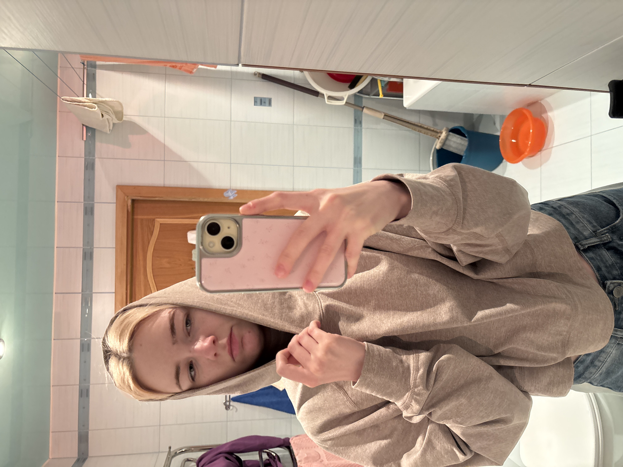 A young woman with blonde hair, wearing a beige hoodie and blue jeans, takes a selfie in a bathroom. She is standing in front of a tiled wall and a mirror. Her left hand is holding the hood of her sweatshirt, and her right hand is holding a pink phone. In the background, a door, a towel rack with towels, a broom, a bucket, and an orange bowl are visible. The overall environment appears to be a domestic bathroom. The location is Unknown, Russia.