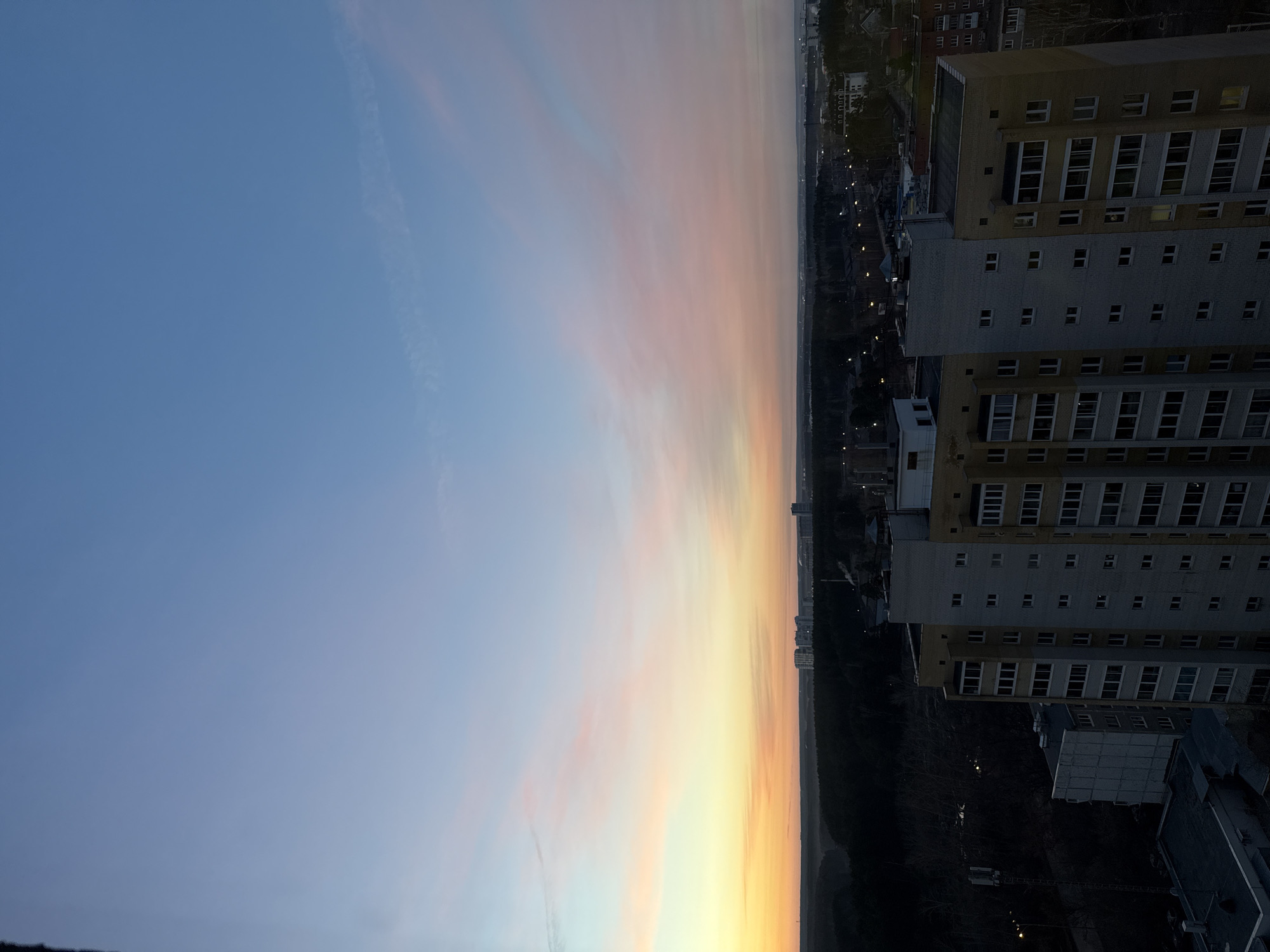 The image shows a high-angle view of Yekaterinburg, Russia, likely taken from a tall building. The sky transitions from blue to orange and yellow hues, indicative of sunrise or sunset. The horizon is dimly lit, with the silhouetted outline of distant buildings and trees. A tall residential building with multiple floors and windows is visible in the foreground, alongside parts of other buildings. The overall impression is that of an urban landscape during twilight.