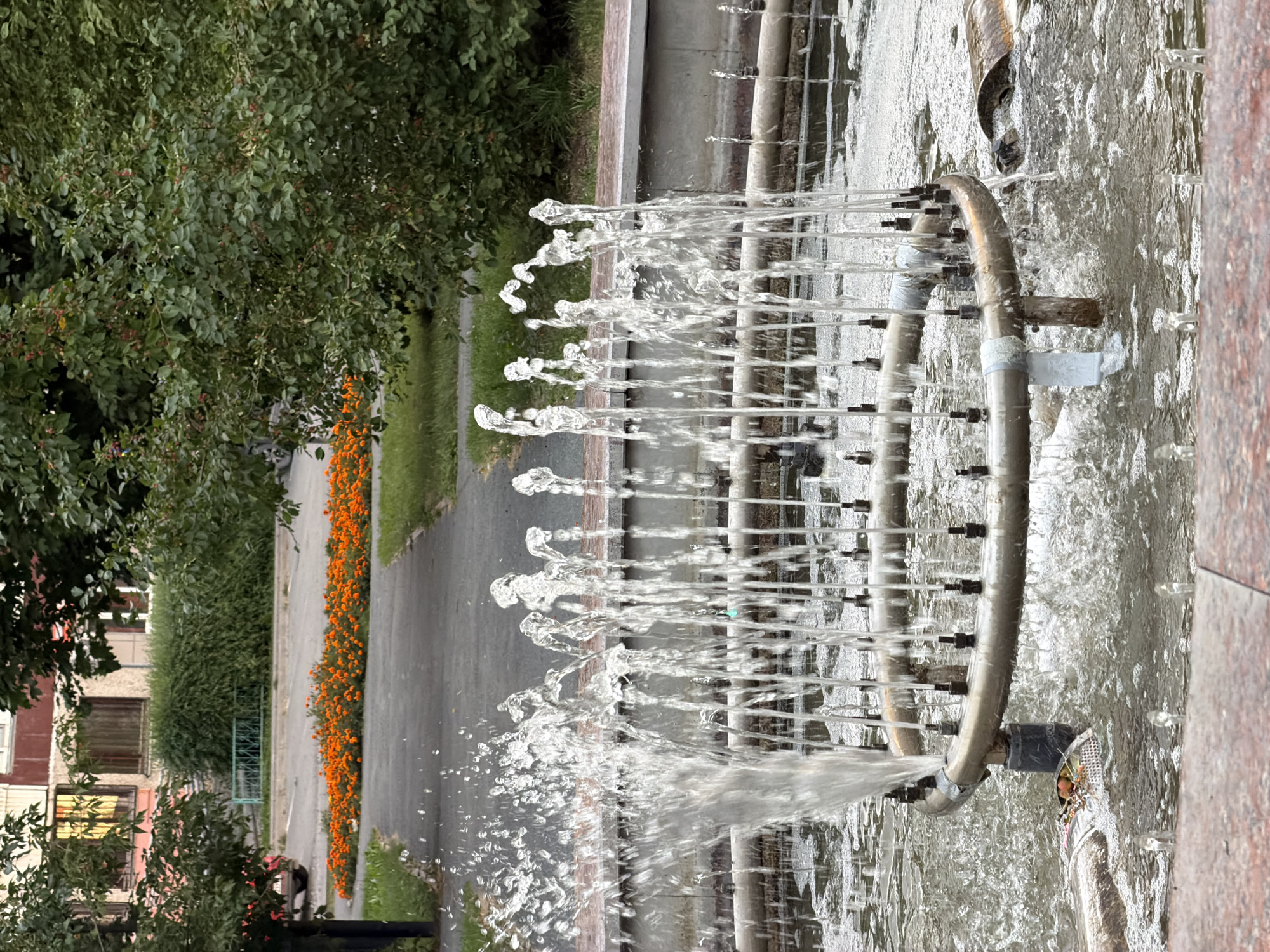 The image shows a close-up of a fountain with water jets. In the background, a pathway leads past a bed of orange flowers, a building, and trees. The fountain appears to be in an urban setting, potentially a park or public space. The overall lighting suggests it is daytime.