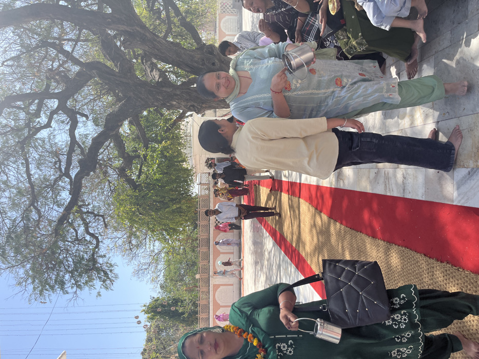 The image shows a group of people standing and sitting on a red and beige carpet in front of a large tree. The carpet is laid out in a path that leads toward a large, white building in the background. The people are dressed in various traditional South Asian clothing. There is a woman in the foreground, wearing a green shirt, holding a black bag in her right hand and a small metal container in her left hand. Next to her, a woman is standing, wearing a beige top and blue jeans. Behind her, a woman wearing a blue and green outfit is handing a small metal container to another woman sitting on the floor. There are several other people standing and sitting in the background. The location of the image is Sector 12, Panchkula, Haryana, India.  The image is taken outdoors, with a sunny sky in the background.