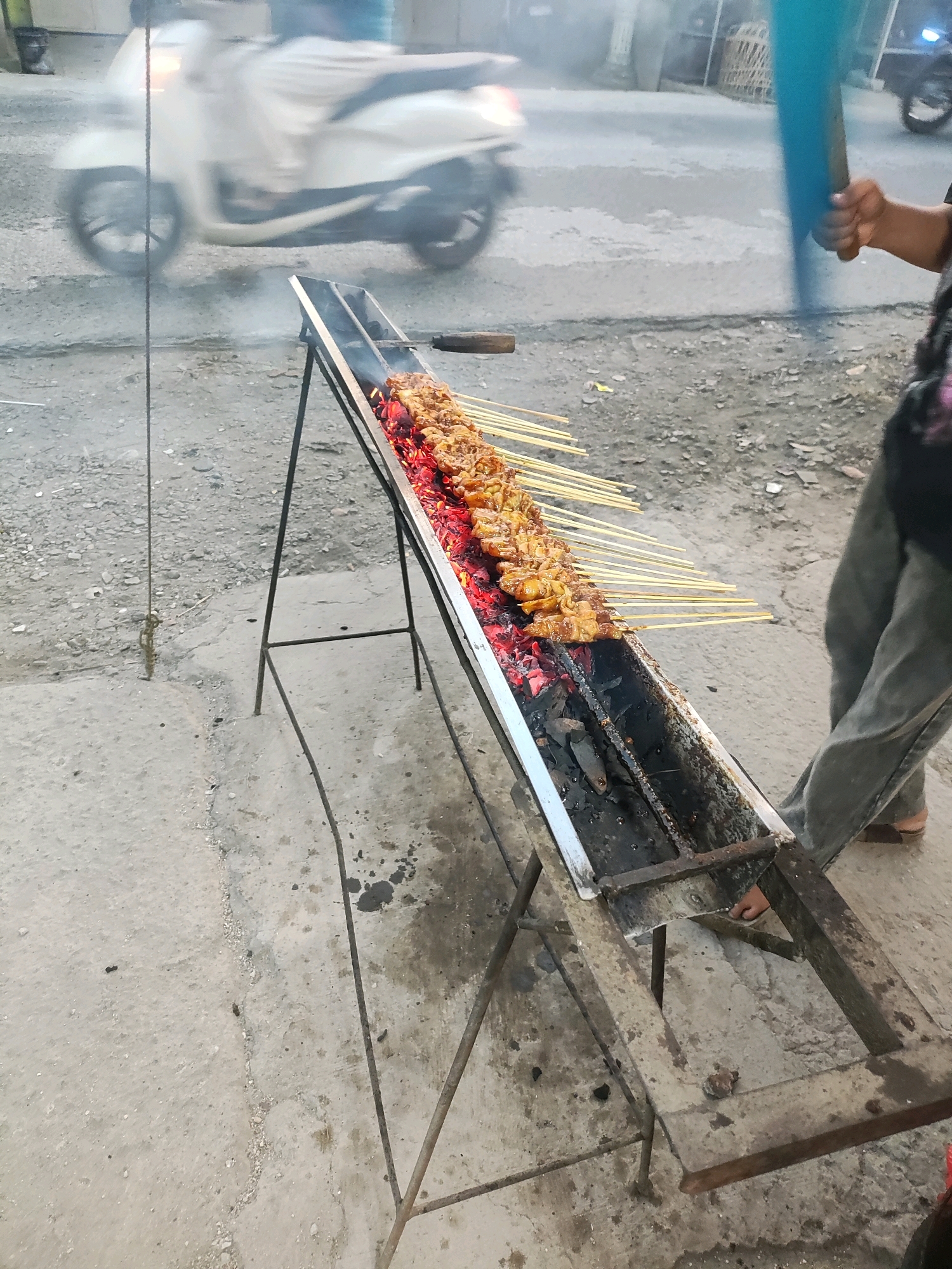 An outdoor food stall in Wanasari, Indonesia, features a long, metal grill filled with glowing red coals, over which skewers of chicken satay are being cooked. The satay is positioned at an angle, with the meat pieces browning and appearing glazed. A person, partially visible on the right, is tending to the grill, holding a large blue object, possibly a fan or a tool for fanning the coals. In the background, a white scooter is in motion, indicating a street setting. The ground is made of rough concrete, and there are signs of wear and tear on the grill and its stand. The atmosphere suggests a busy street food environment.