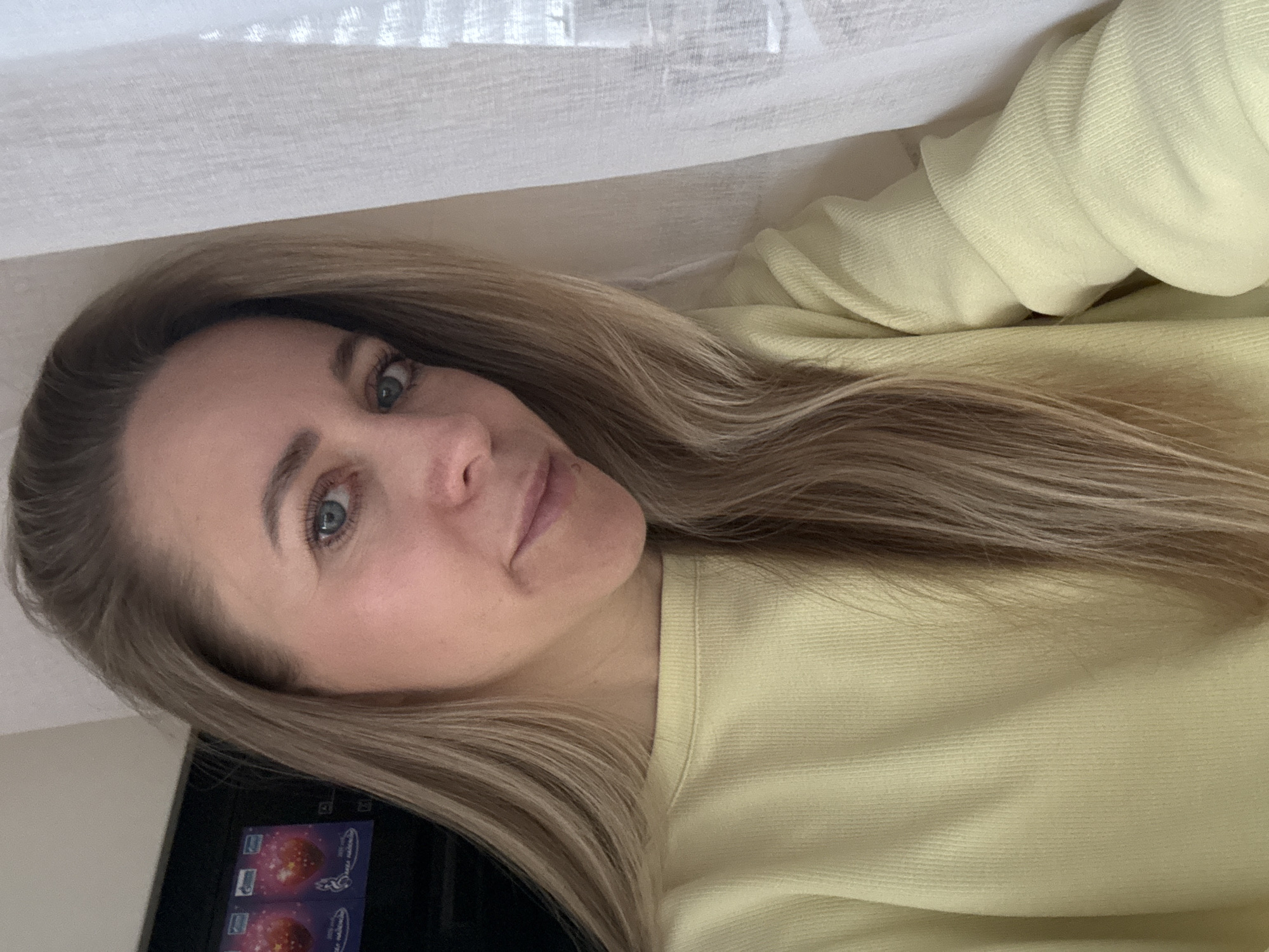 A woman with light blonde hair and blue eyes is facing the camera. She is wearing a light yellow sweater. A white fabric is draped above her. In the background, there is a black object with a colorful sticker on it. The location cannot be determined from the provided image.