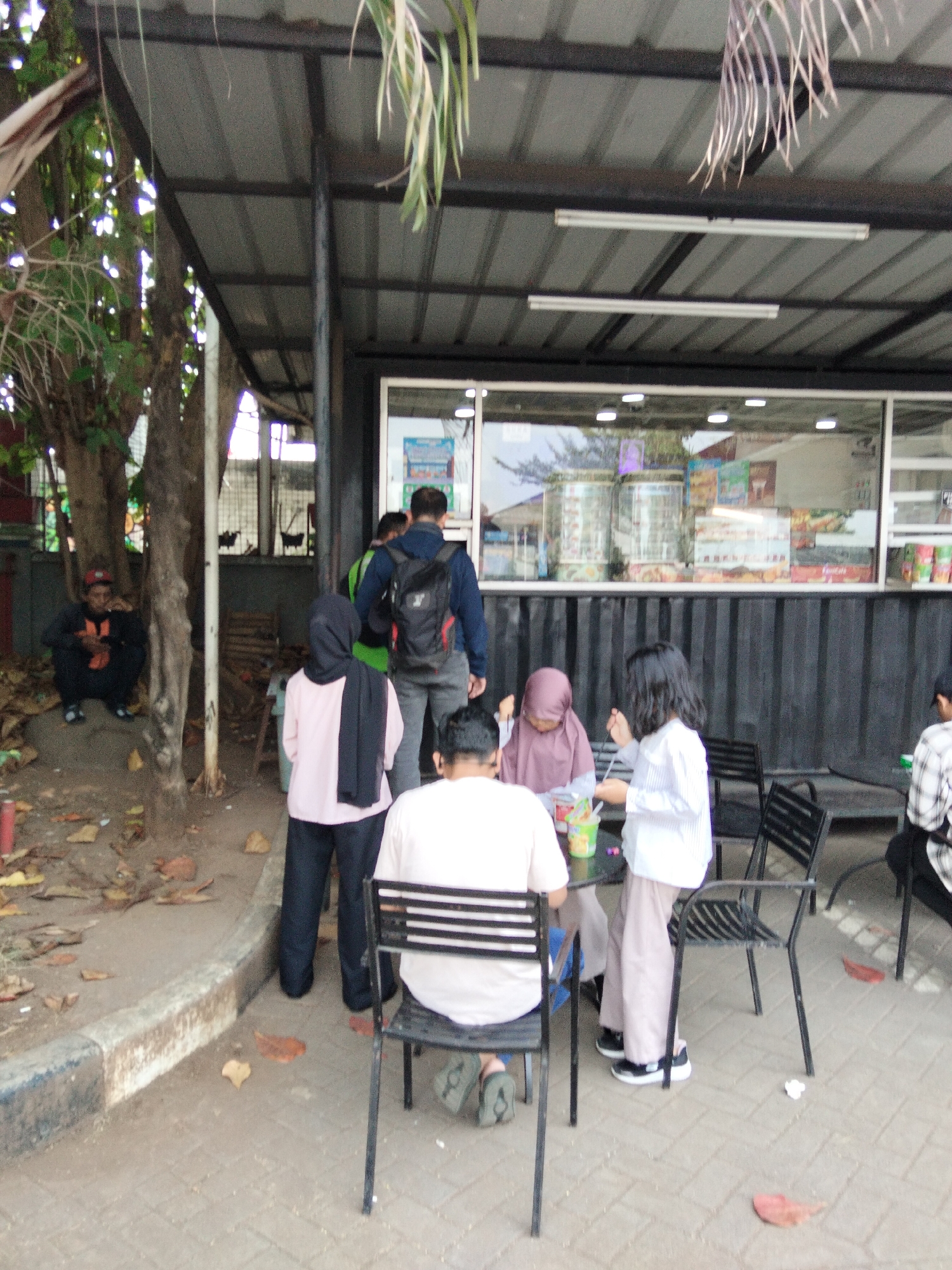 In the city of South Tangerang, Indonesia, a group of people are gathered outdoors at a food stall or cafe. The scene depicts an open-air seating area with several tables and chairs. In the foreground, a person is seated facing away from the camera, wearing a light-colored shirt and dark pants, with their legs crossed as they sit at a black metal chair. To their left, another person in a light pink shirt and dark pants, wearing a black headscarf, stands and appears to be interacting with someone at the table.
Behind them, a person wearing a purple headscarf and a light-colored top is seated at the table, looking down. A woman in a white striped shirt and light-colored pants stands to the right of the table, actively engaged with the group. Several cups and a bottle are visible on the table.
Further back, a person in a dark jacket and backpack is walking away, while another person wearing a green shirt and a dark headscarf stands near a tree. To the far left, a person in an orange and black outfit is crouched near a tree trunk.
The environment includes a corrugated metal roof supported by metal poles, a large window or glass display with items visible inside the stall, and a textured paved ground. Fallen leaves are scattered on the ground, suggesting an outdoor setting. The overall impression is a casual gathering at an eatery.
