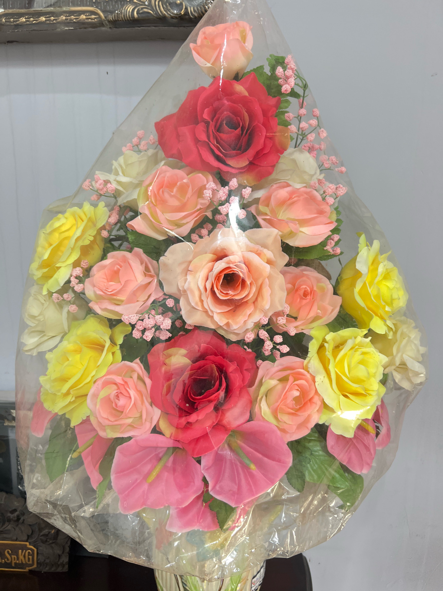 A bouquet of artificial flowers is displayed, encased in clear plastic. The bouquet features a variety of roses in shades of red, pink, and yellow, interspersed with smaller pink filler flowers and some greenery. Additionally, there are pink anthurium flowers visible towards the bottom of the arrangement. The bouquet is positioned against a plain, light-colored wall, possibly a home interior. In the lower left corner, a portion of a dark object with a brass plate bearing the inscription "SP.KG" is visible. This scene does not provide any specific visual cues to pinpoint its exact location within Denpasar, Indonesia, or any other specific city.