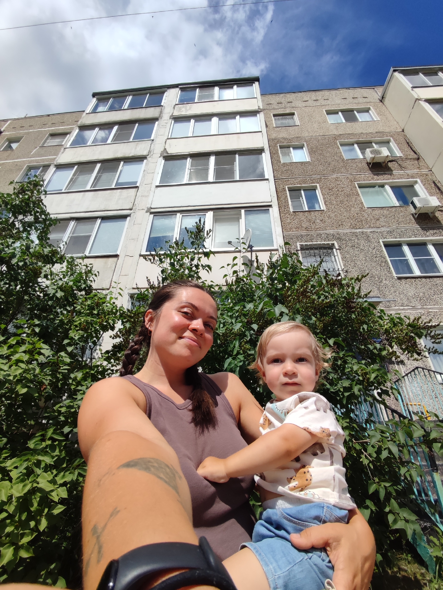 A woman and a young child are pictured from a low angle, looking up towards a multi-story apartment building under a partly cloudy sky. The building is constructed of gray concrete with a textured finish and features numerous windows. Some windows are covered with blinds, while others reflect the sky. Air conditioning units are visible on some of the building's facades. Green foliage, likely bushes or small trees, surrounds the lower part of the building and obscures some of its base. A metal railing can be seen on the right side of the frame, suggesting a balcony or stairway. The woman, wearing a dark tank top, has dark hair in a braid and is holding the child. She has a tattoo on her left arm and is wearing a watch on her left wrist. The child, with blonde hair, is wearing a light-colored t-shirt with animal prints and denim shorts. The child is looking directly at the camera with a neutral expression. The woman is smiling slightly. The image appears to have been taken during the daytime, likely late morning or afternoon, given the brightness of the sunlight and the blue sky. The presence of the apartment building and the foliage suggests an urban or suburban residential setting. The overall mood is casual and familial, with the subjects posing for a selfie. There is no visible text in the image.