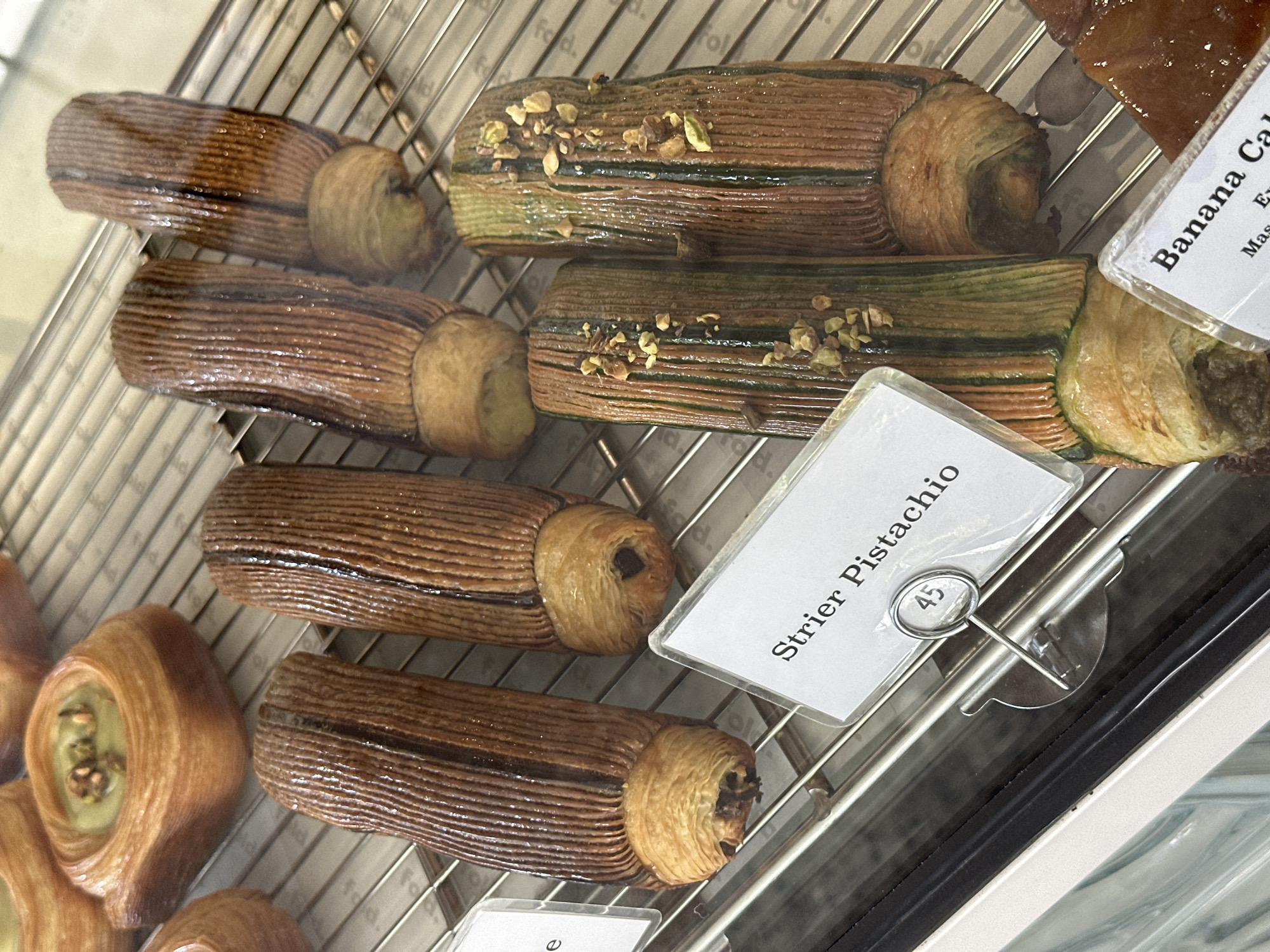 The image shows a selection of pastries displayed on a wire rack, likely in a bakery or cafe. The sign indicates that one of the pastries is "Strier Pistachio," priced at 45, presumably Indonesian Rupiah (IDR). The pastries appear to be laminated dough, with some topped with what looks like chopped pistachios. A sign for "Banana Cake" is also visible. The scene likely takes place in the Ubud District, Indonesia, based on the implied currency. The lighting appears to be indoor and artificial, highlighting the pastries' glossy glaze.