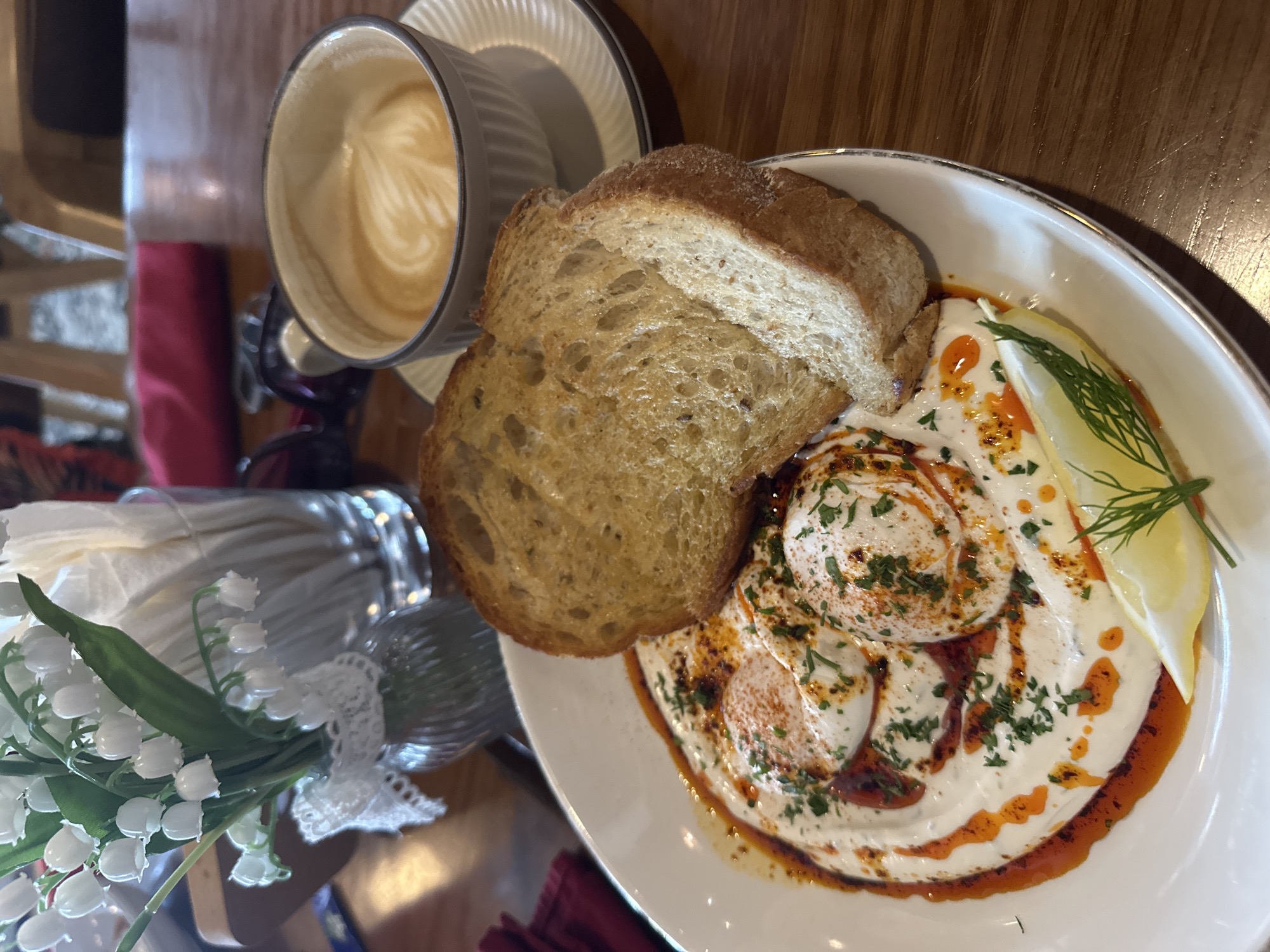 A high-angle shot captures a breakfast setting on a wooden table. A white plate contains what appears to be poached eggs nestled in a swirl of yogurt or cream sauce, drizzled with chili oil and garnished with parsley and dill. Beside the eggs, two slices of toasted bread rest against the plate. Adjacent to the plate, a cup of latte sits on a saucer. To the left, a glass vase holds a bouquet of white flowers. The setting resembles a cafe or restaurant. There is no indication of it being in Nha Trang Ward, Vietnam.