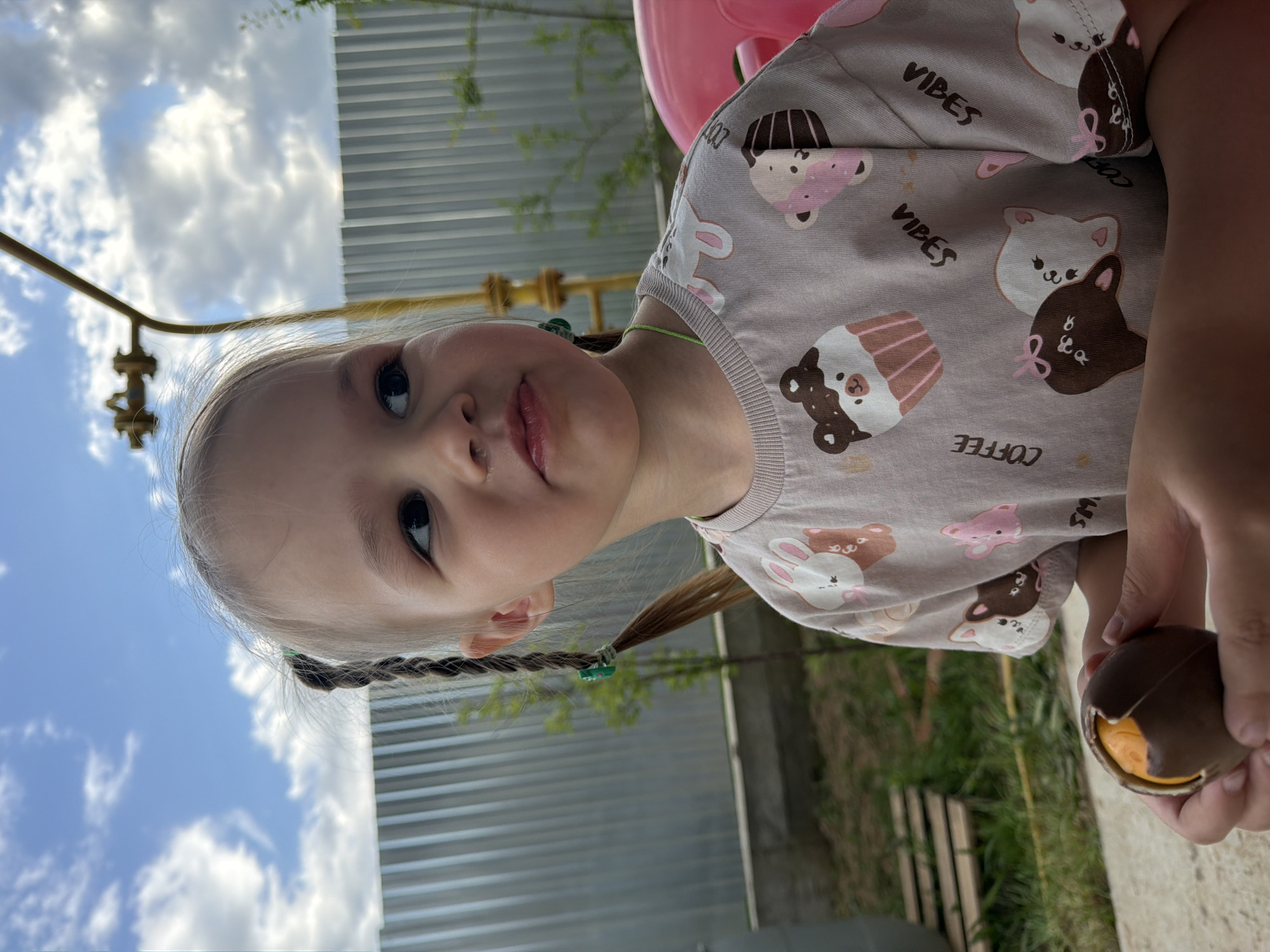 A young child with braided hair, wearing a t-shirt with cartoon animal and cupcake designs and the word "VIBES," is holding a chocolate candy. The photo appears to be taken outdoors near a playground or park, with a glimpse of a cloudy sky and what seems to be a metal fence in the background. The photo might be taken in the city of Chaikovsky, Russia, though specific location confirmation is impossible without further information.