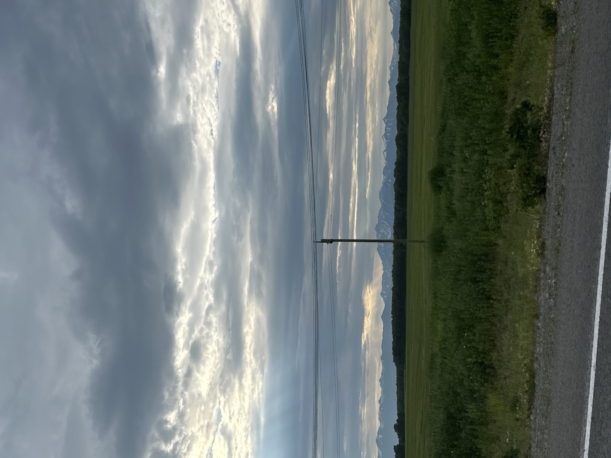 The image depicts a rural landscape under a cloudy sky with sunlight filtering through in some areas. In the foreground, a paved road with a white line is visible. Beyond the road is a green field with tall grass and some smaller plants. In the mid-ground, a dark line of trees stretches across the horizon, backed by a range of snow-capped mountains. A single wooden utility pole stands in the field, with wires stretching horizontally across the frame. The scene appears to be set in the city of Кеткино, Russia.