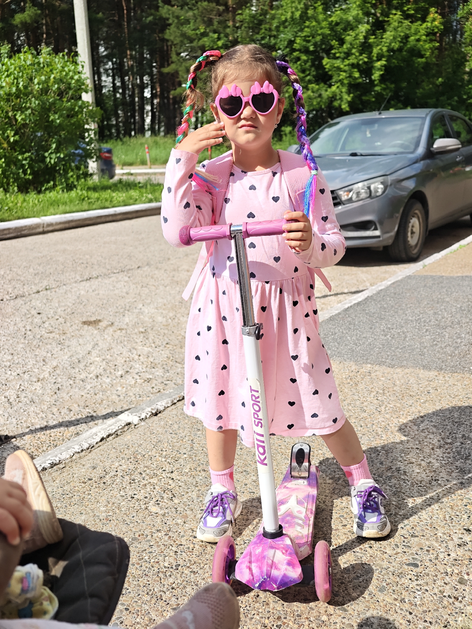 A young girl with pink heart-shaped sunglasses and colorful braided hair stands on a sidewalk in Chaikovsky, Russia, next to a pink and white scooter. She is wearing a pink long-sleeved dress adorned with black hearts and purple shoelaces on her white sneakers. The girl is looking towards the camera, her left hand touching her face. In the background, a gray sedan is parked on the street, and trees line the area. The pavement is textured with small pebbles. A stroller is partially visible in the lower left corner of the frame.