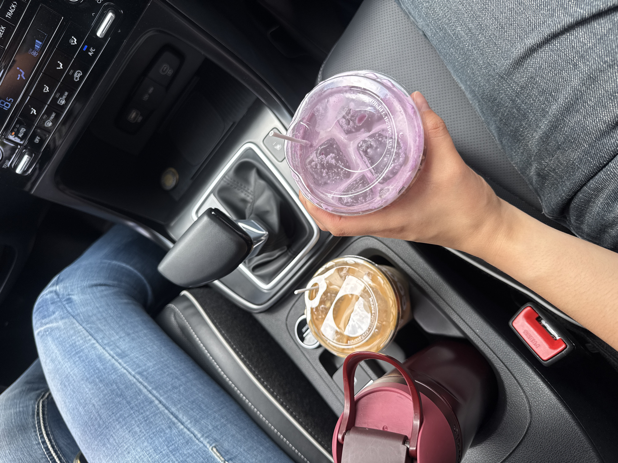 This image captures the interior of a car, likely in Puebla, Mexico, from a slightly elevated, top-down perspective. A person, identifiable by their legs clad in blue jeans and a visible arm and hand, is seated, possibly in the passenger seat.
The main activity shows the person holding a clear plastic cup filled with a light purple beverage, containing ice and a white straw, suggesting they are either enjoying a drink while stopped or during a journey. In the car's center console, two other beverages are visible in cup holders: another clear plastic cup containing iced coffee with a straw, and a dark red or maroon water bottle. To the right of the cup holders, a red seatbelt buckle is prominent, with the word "PRESS" clearly visible on it.
The car's dashboard and center stack are visible on the left side of the frame. The climate control panel features a digital display showing "18.5" (likely temperature), and several buttons with visible text including "TEMP", "AUTO", "LOW", "OFF", "FRONIT", "REXR", and "A/C". Above these controls, a partial label "EK TRACK>" can be seen. Below the main controls, a compartment reveals a USB port and a 12V power socket. The interior appears clean and modern, with black and grey textures. The overall lighting suggests it is daytime.