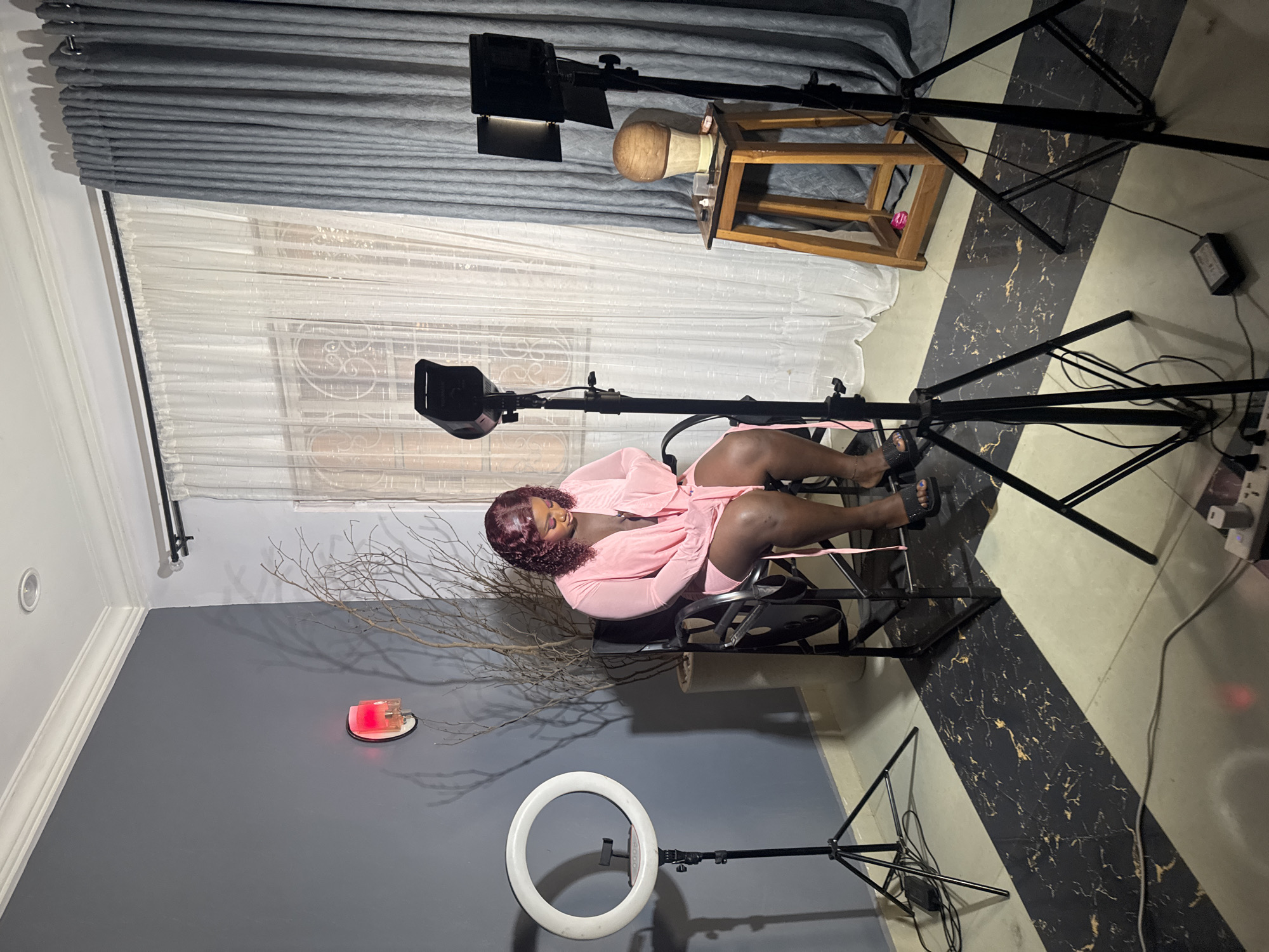 A Black woman is seated in a director's chair in what appears to be a home studio setup in Gura Top, Nigeria. She is wearing a pink robe and has curly, dark hair. In front of her, to her left, is a ring light on a tripod. To her right, there are two studio lights on tripods and a wooden stool with a wig on a stand. The room has gray walls and the floor is tiled with a black and gold pattern. She is looking down at her hands.