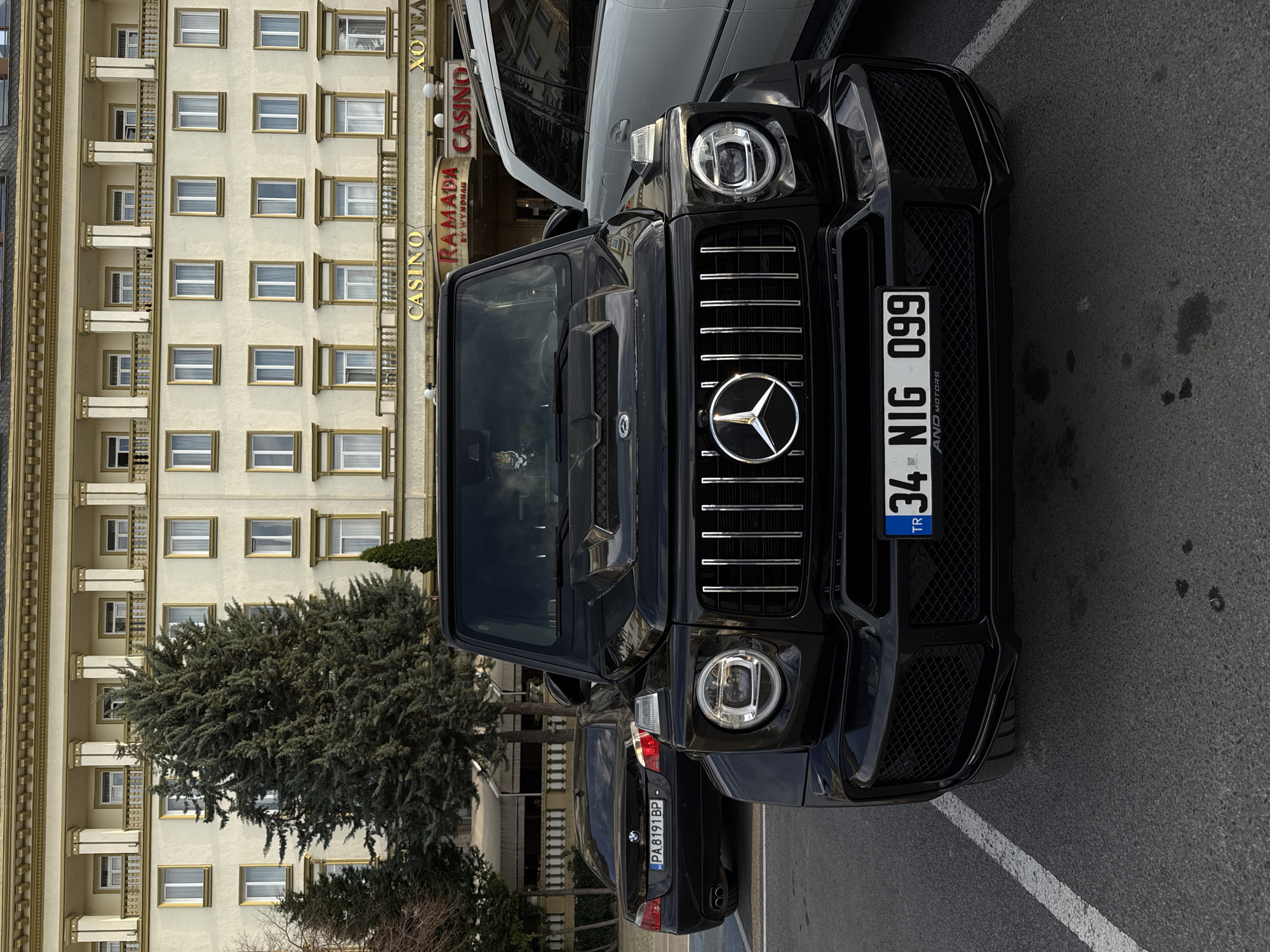 The image shows a black Mercedes-Benz G-Class SUV parked on a street in Plovdiv, Bulgaria. The license plate reads "34 NIG 099," indicating that the car is registered in Turkey. Behind the SUV, another car with license plate "PA 8191 BP" is visible. In the background is the Ramada by Wyndham Plovdiv Trimontium Hotel, featuring its "CASINO" sign. A large tree stands in front of the hotel, partially obscuring its facade. The street appears to be marked with white parking lines. The angle of the photograph presents the scene with a slight sideways tilt.