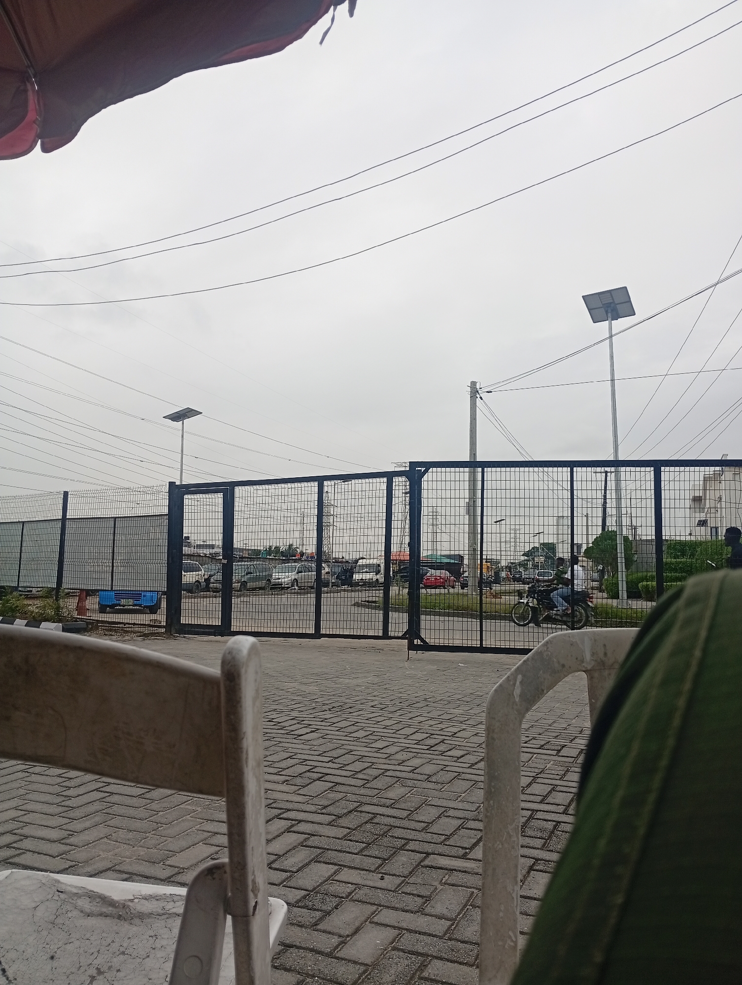 The image depicts a street scene in Ilado, Nigeria, under an overcast sky. In the foreground, a portion of a green fabric object is visible on the right, and a white plastic chair is partially seen on the left. Beyond the chairs, a paved walkway made of interlocking gray bricks leads to a large black metal gate. Through the gate, a parking lot or open area is visible, containing several vehicles including cars and a motorcycle with two riders. In the background, power lines crisscross the sky, and solar-powered street lights are present. A security fence runs along the perimeter of the area behind the gate. The overall impression is of a somewhat industrial or commercial area on a cloudy day.