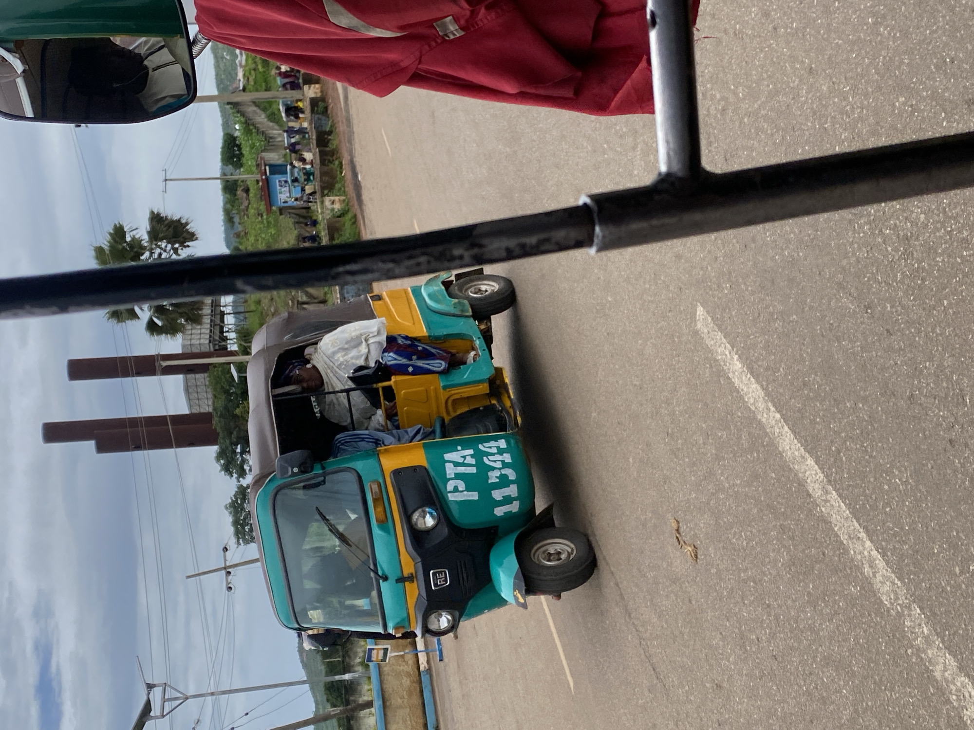 The image shows a turquoise and yellow motorized rickshaw, commonly known as an auto-rickshaw or "keke napep", on a paved road. The vehicle has "PTA 11344" written in white letters on its side. A woman in a white garment and headscarf is seated in the passenger area of the rickshaw. The rickshaw is moving, as indicated by the motion blur of the wheels. In the background, there are industrial chimneys and some vegetation, suggesting an outdoor, possibly semi-urban or rural setting. The sky is overcast. The foreground shows a metallic structure, possibly part of another vehicle or a frame, and a red garment. The image is likely taken in Rafin Par, Nigeria, given the presence of this common mode of transportation and the visual cues of the environment.