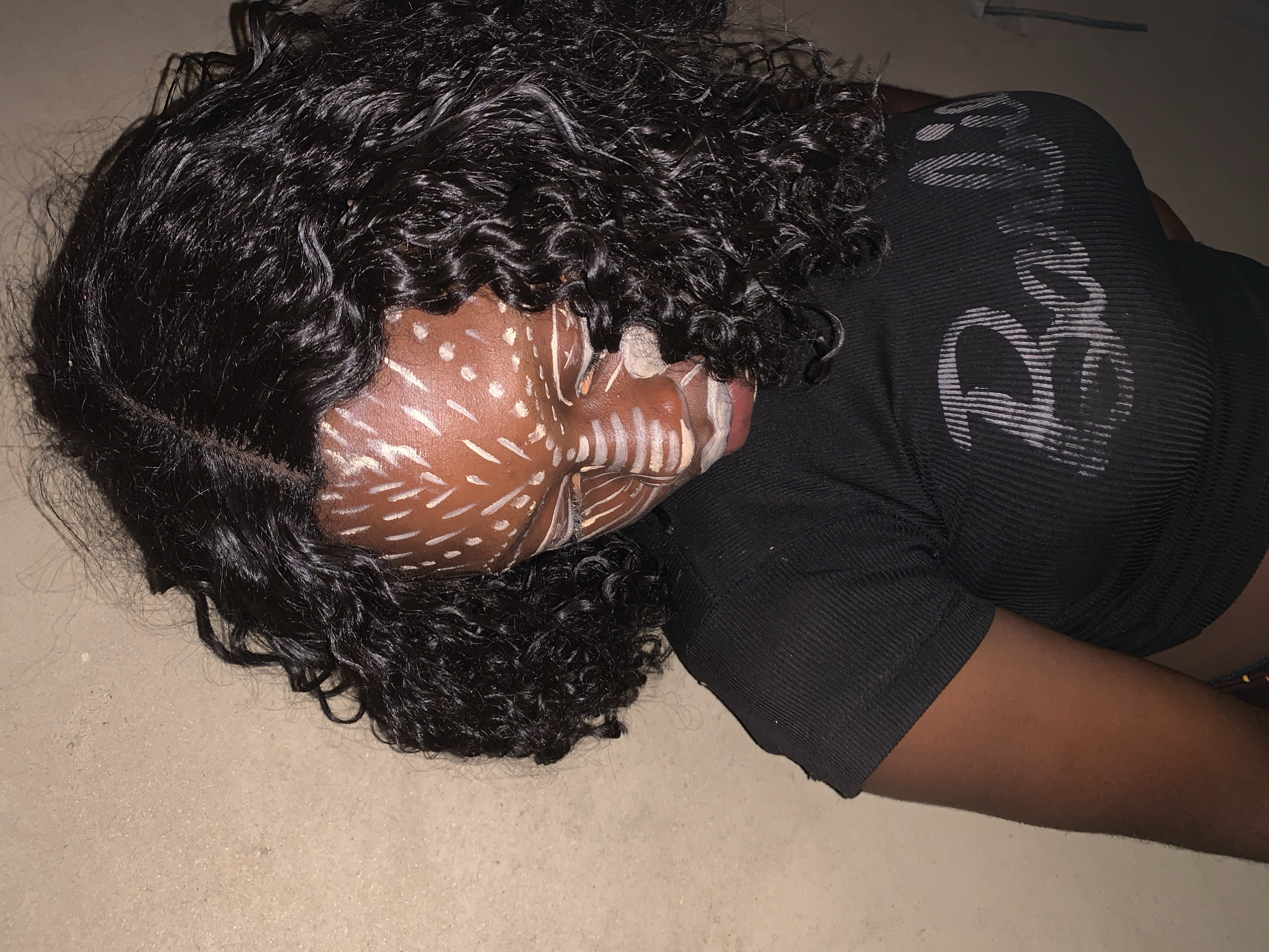 A person with dark curly hair, wearing a dark-colored top, lies on a light-colored surface. Their face is painted with white markings. The location is unknown, and there is no information to suggest this is the city of Agban, Nigeria.