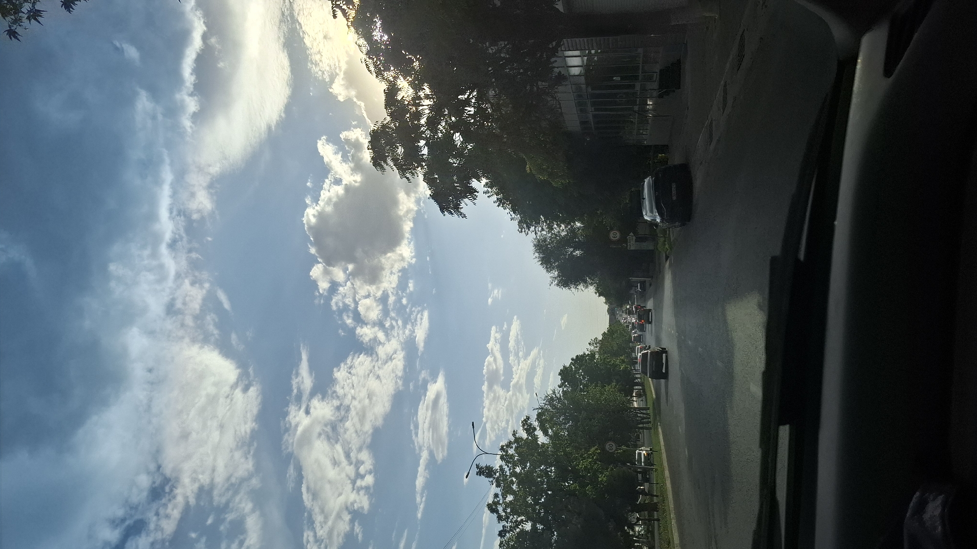 The image depicts a street view in Shymkent, Kazakhstan, as seen from inside a vehicle. The scene is dominated by a wide, paved road with multiple lanes, bordered by trees and buildings. Several cars are visible on the road, indicating moderate traffic. The sky above is a mix of blue and white clouds, with sunlight casting a warm glow, suggesting it is either morning or late afternoon. The trees lining the street are lush and green, indicating a warm season. A speed limit sign of "60" is visible on the right side of the road. The foreground shows the dashboard and windshield of the vehicle from which the photo was taken. The overall impression is a typical urban street scene during daylight hours.