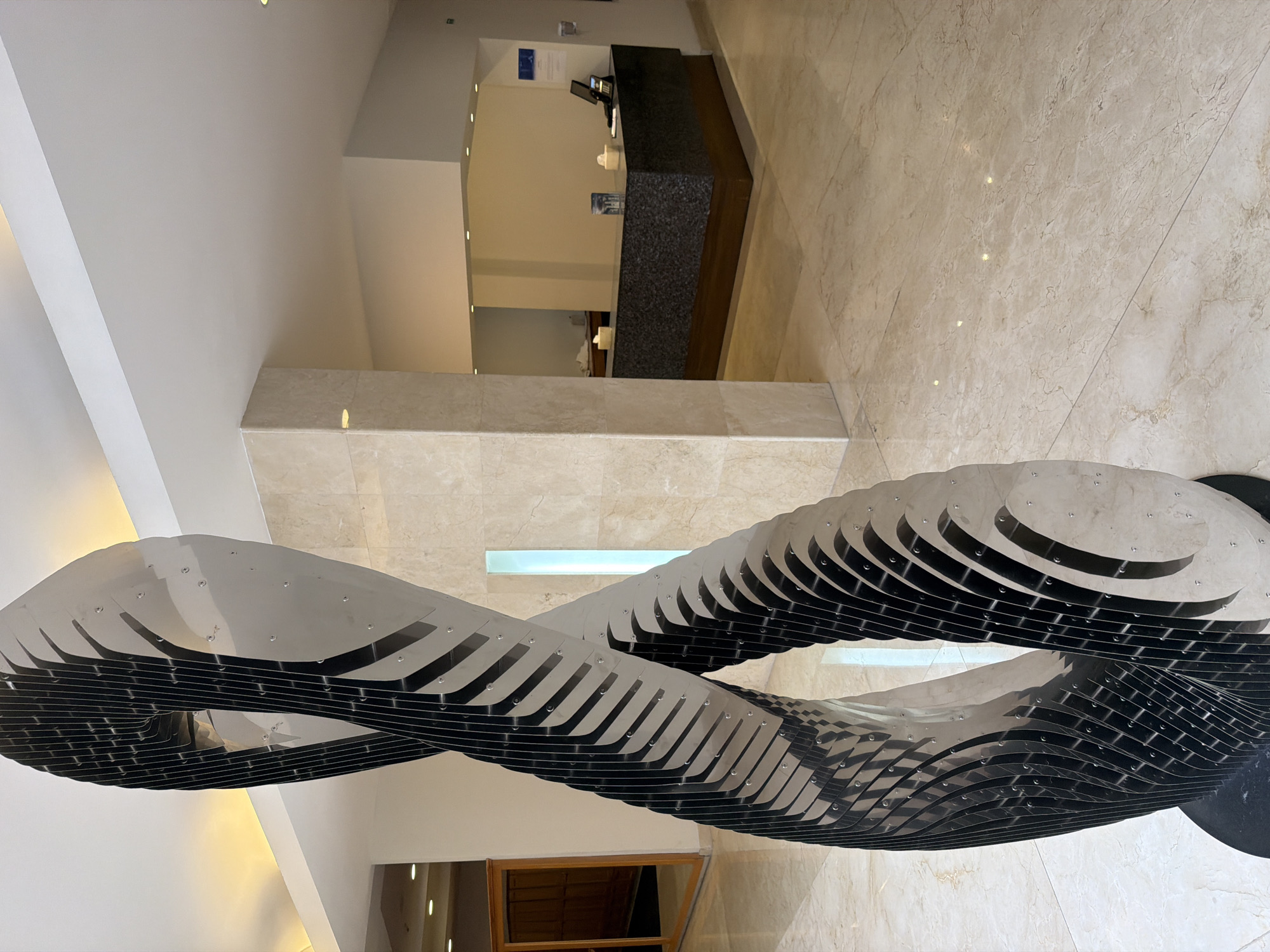 The image captures the interior of a building in San Bernardino Tlaxcalancingo, Mexico. The foreground features a large, modern art sculpture made of layered, metal slices forming an abstract, twisting shape. The floor is tiled in a light beige marble pattern. In the background, there is a reception desk with a dark counter and light-colored wall behind it. The lighting appears to be a mix of natural and artificial. There are no people visible in the image.