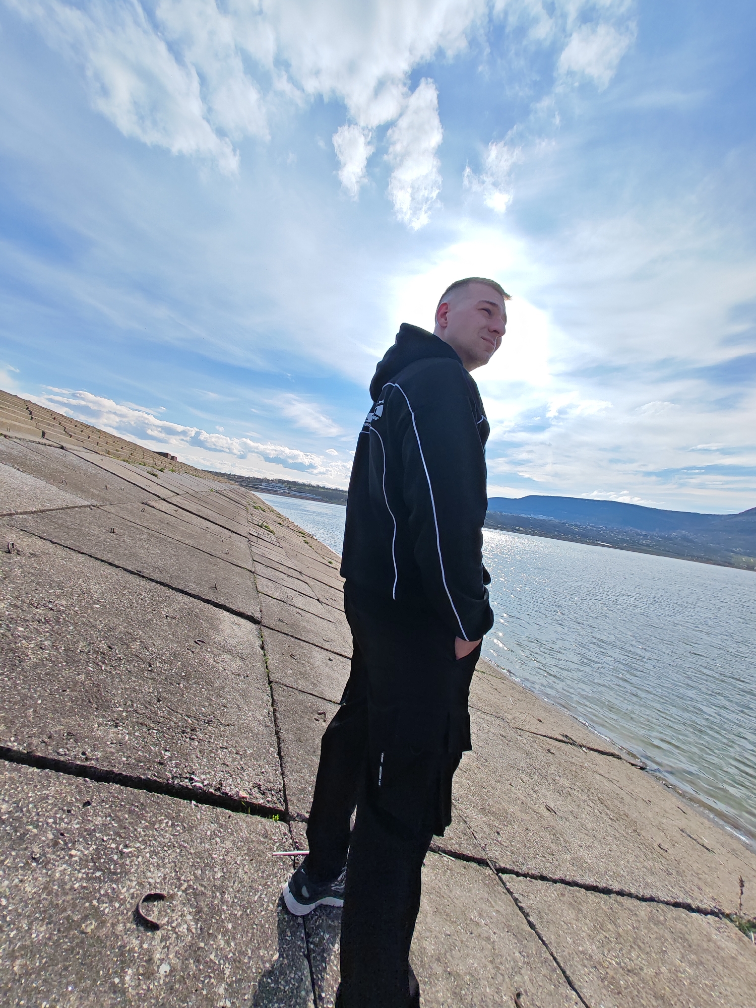 A young man stands on a concrete incline next to a body of water under a partly cloudy sky with the sun shining. He is facing away from the viewer, looking to his right. He is wearing a black hooded jacket with white trim and black cargo pants. He has short blonde hair. The concrete incline has cracks and embedded metal hooks. Beyond the water, a line of hills and a few buildings are visible. The image was taken in the city of Shumen, Bulgaria.