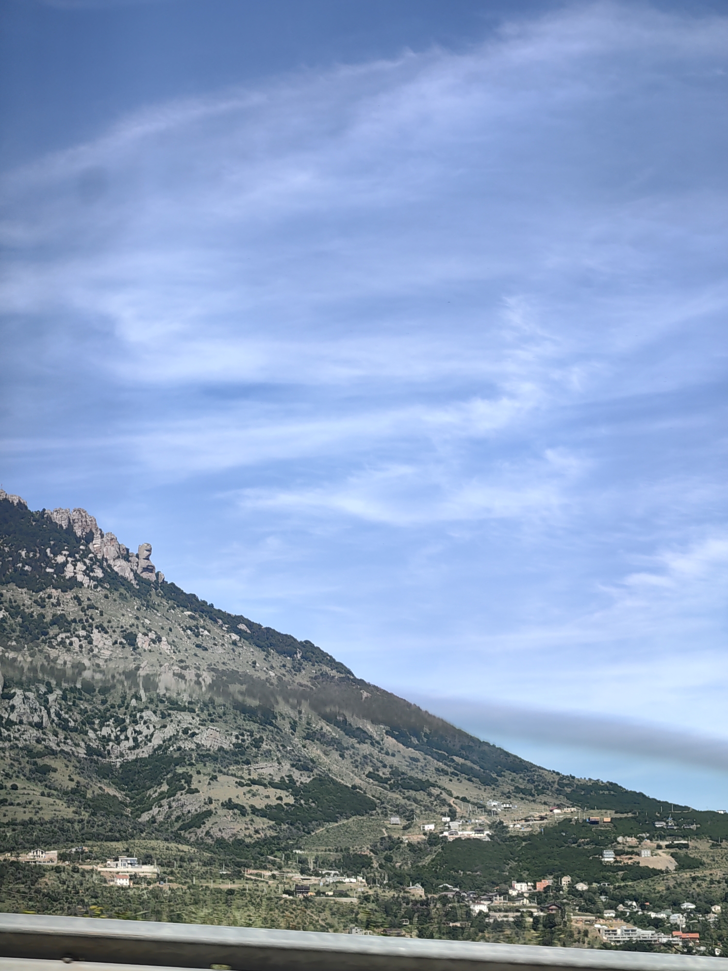 The image shows a mountainous landscape under a partly cloudy blue sky. The mountains are covered with sparse vegetation and rocky outcrops. In the mid-ground, there is a cluster of buildings, suggesting a settlement. A metal barrier is visible in the foreground, running horizontally across the bottom of the image. The scene appears to be a scenic view of the outskirts of Kiparisnoe, Ukraine, with the mountains forming the backdrop to the residential area.