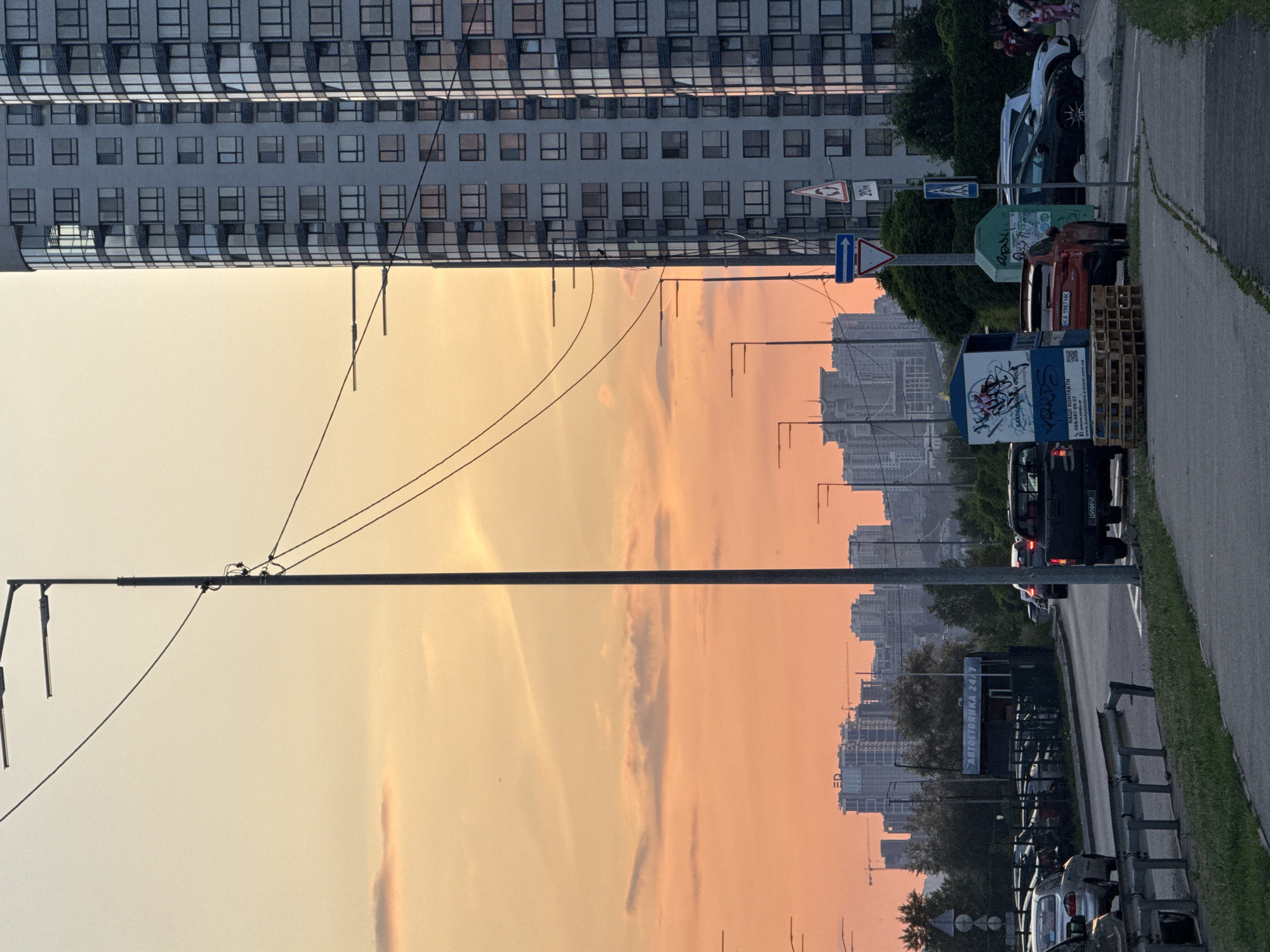 The image depicts an urban street scene at sunset or sunrise, characterized by a warm, orange and yellow-tinged sky that dominates the central portion of the frame. The perspective is unusual, looking across the sky between a tall, modern building and a busy street.
On the left, a multi-story building with numerous windows reflects the soft light, showcasing a contemporary architectural style with both flat and curved sections. Utility poles with overhead wires run across the image, silhouetted against the bright sky.
To the right, a road or street is visible with several parked and possibly moving vehicles. Cars of various models and colors are lined up along the curb. Small figures, likely people, are faintly visible on the sidewalk in the background, next to some of the parked cars. Along the roadside, there are grassy patches, trees, and street furniture, including metal fences.
Notable objects include several traffic signs: an inverted red triangle (Give Way), a blue pedestrian crossing sign, a sign indicating "20m", and a blue arrow sign. An advertising stand or kiosk with graffiti and Cyrillic text is present, along with a green container also covered in graffiti. Further down the street, another stand clearly displays "АВТОСТОЯНКА 24/7" (Parking 24/7) and "НАШІ КОНТАКТИ" (Our Contacts) with a phone number and website "o.kram.com.ua". A red car's license plate "KA 1987 HK" and a black van with "ЦИФРИ" (Numbers) on its rear are visible.
In the far background, a hazy city skyline with more buildings and several construction cranes suggests ongoing development. The overall impression is an everyday urban environment bathed in the dramatic light of late evening or early morning, set in Kyiv, Ukraine, as indicated by the Cyrillic text and license plate format.