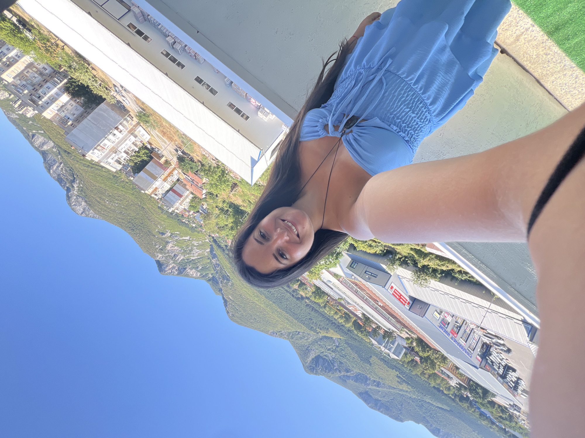 A woman with long dark hair and a light blue strapless dress stands on a balcony, taking a selfie. She is smiling at the camera. Behind her, a city with apartment buildings and a large, long industrial building with a white roof is visible, nestled against a backdrop of green mountains under a clear blue sky. The scene appears to be the city of Vratsa, Bulgaria.