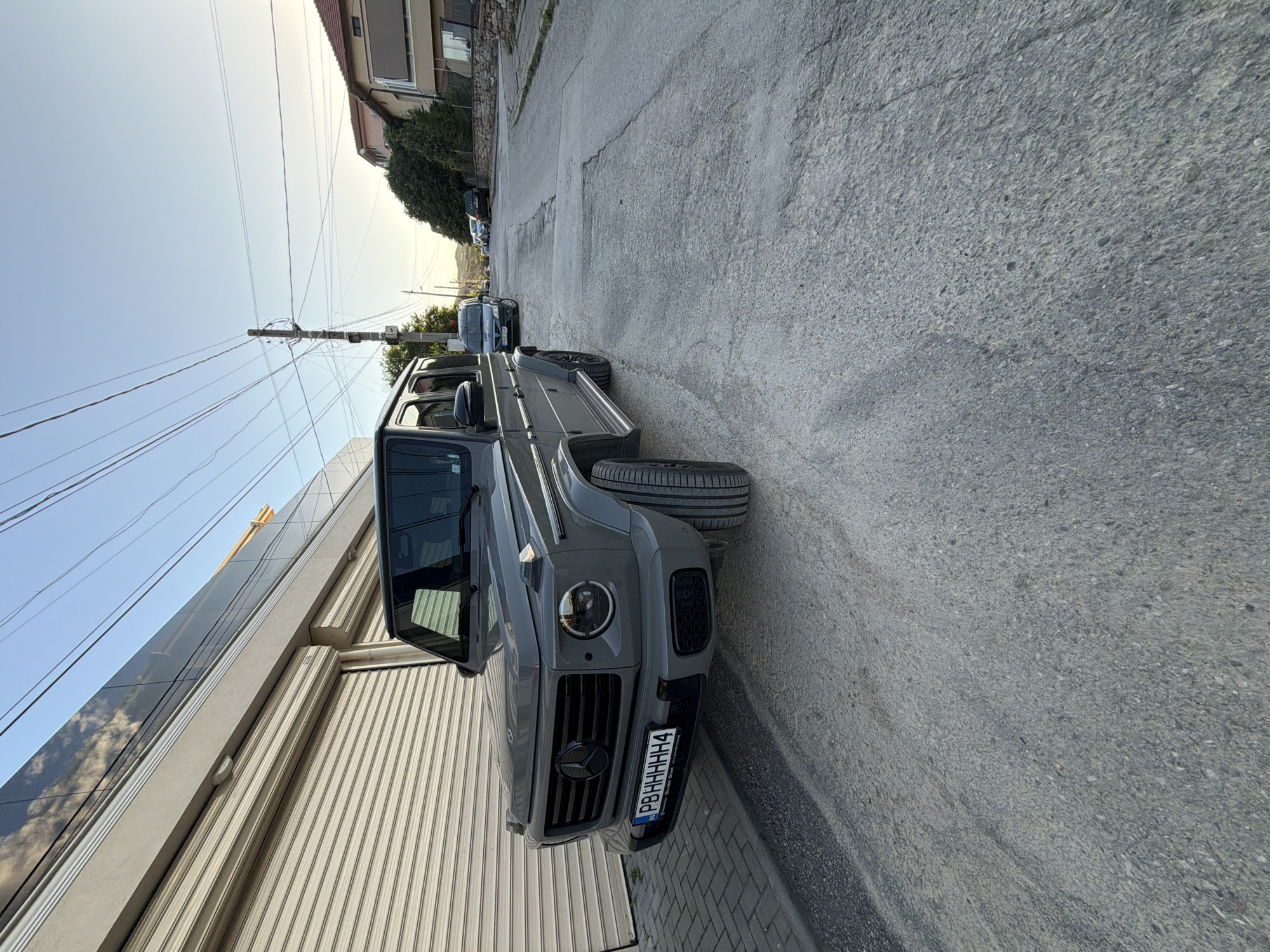The image shows a gray Mercedes-Benz G-Class (G-Wagon) parked on the side of a narrow street in Asenovgrad, Bulgaria. The vehicle is facing the camera. The license plate is visible and reads "PBHHHHHH4".
On the left, there is a modern building with a corrugated metal exterior and reflective windows. Overhead, there are power lines crossing the sky.
In the background, other parked cars are visible down the street, leading to a residential area. The street surface is asphalt with visible cracks and patches. The sky is clear and bright, suggesting it is daytime. The image appears to have been taken from a low angle.