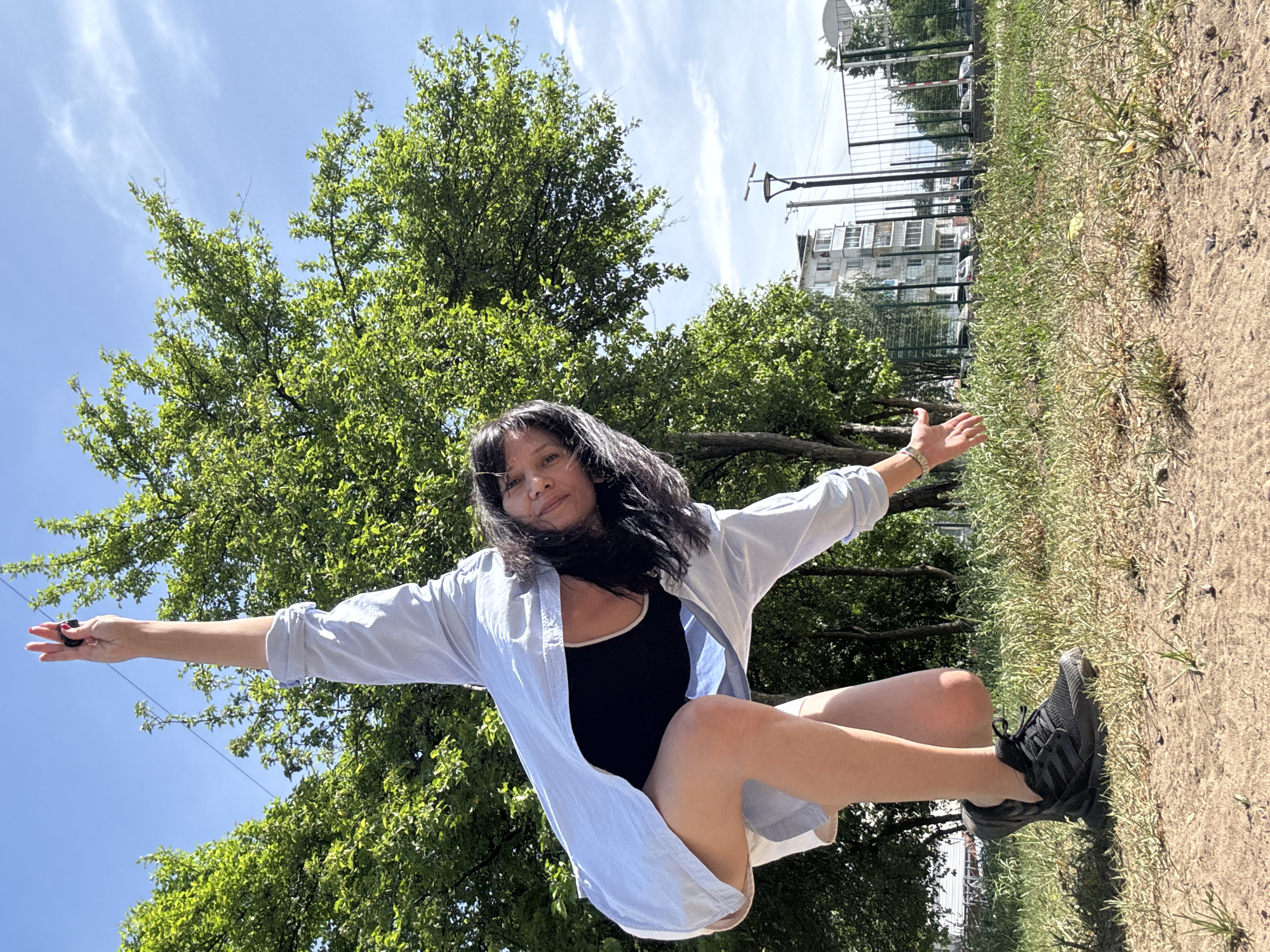 A full shot of a woman in a black tank top and light blue button-down shirt posing in a park on a sunny day in Unknown, Unknown. The woman is kneeling with her arms outstretched on a grassy patch of land. She wears black sneakers. A large tree stands behind her, with a blue sky and white clouds visible above. In the background, there is a fence, a building, and a basketball hoop. The ground is partly grassy, partly sandy.
