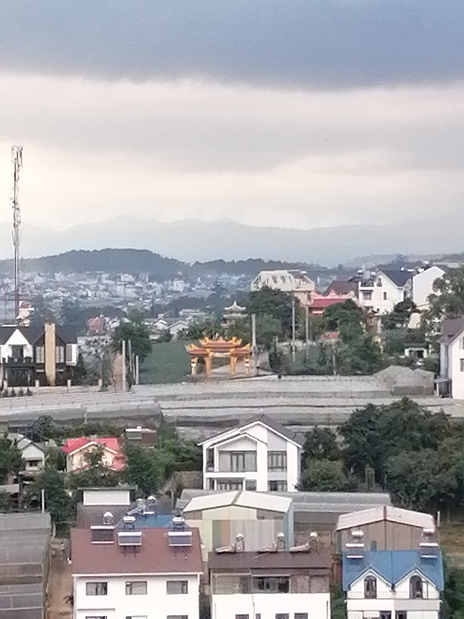The image displays a panoramic view of a residential area in Xuân Trường - Đà Lạt, Vietnam, under an overcast sky. In the foreground, several multi-story houses with various roof colors and architectural styles are visible. Some houses have red or blue roofs, and some have white exteriors. There are also glimpses of trees and some construction materials or infrastructure.
Further into the background, a more densely populated urban landscape unfolds, with numerous buildings of different sizes and designs. A prominent feature in the mid-ground is an ornate, yellow gateway or shrine, possibly part of a temple or cultural site. Beyond the urban sprawl, a range of hills and mountains can be seen, fading into the distance under the cloudy sky. A tall communication tower is visible on the left side of the frame, standing out against the sky and the distant landscape. The overall scene suggests a typical hillside town in Vietnam, characterized by a mix of modern housing, older structures, and natural topography. There are no people clearly visible in the image.