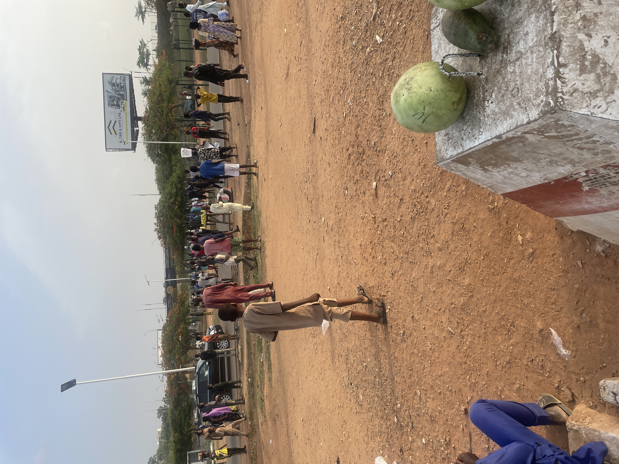 The image depicts a scene in Abuja, Nigeria, characterized by a dusty, brown, dirt-covered ground. A line of people extends into the distance, generally moving from right to left in the image, along the edge of a road. The people are of various heights and clothing styles, suggesting a diverse group. A billboard is visible further up the road. In the foreground to the right, there is a weathered concrete block or post with a white and red stripe, where several green fruits are kept for sale. A boy stands in the right center ground. Some people wearing blue trousers and sandals are seated near the bottom-right. The environment suggests a hot and sunny day.