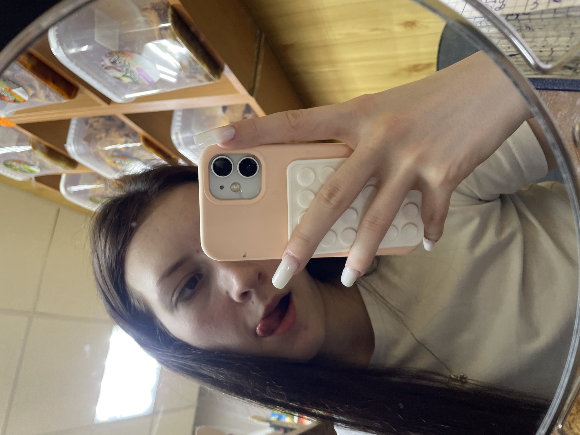 A young person, likely in Semyonov, Russia, captures a mirror selfie, holding a light pink/beige smartphone with a white, circular pop-it style grip on its back. The phone has two prominent camera lenses. The individual has long, dark hair and long, white manicured nails. They are making a playful expression, sticking out their tongue, and looking directly into the camera/mirror.
The reflection in the mirror reveals an indoor setting. Behind the person, there's a wooden shelving unit containing multiple clear plastic containers, possibly holding food items or snacks, with some colorful labels visible inside. Above, a light-colored ceiling with a bright light source is reflected. To the right of the reflection, part of a wooden surface, some papers with faint, unreadable text, and a metallic frame are visible. The person is wearing a light-colored top and a thin necklace with a small pendant. The overall impression is casual and cheerful.