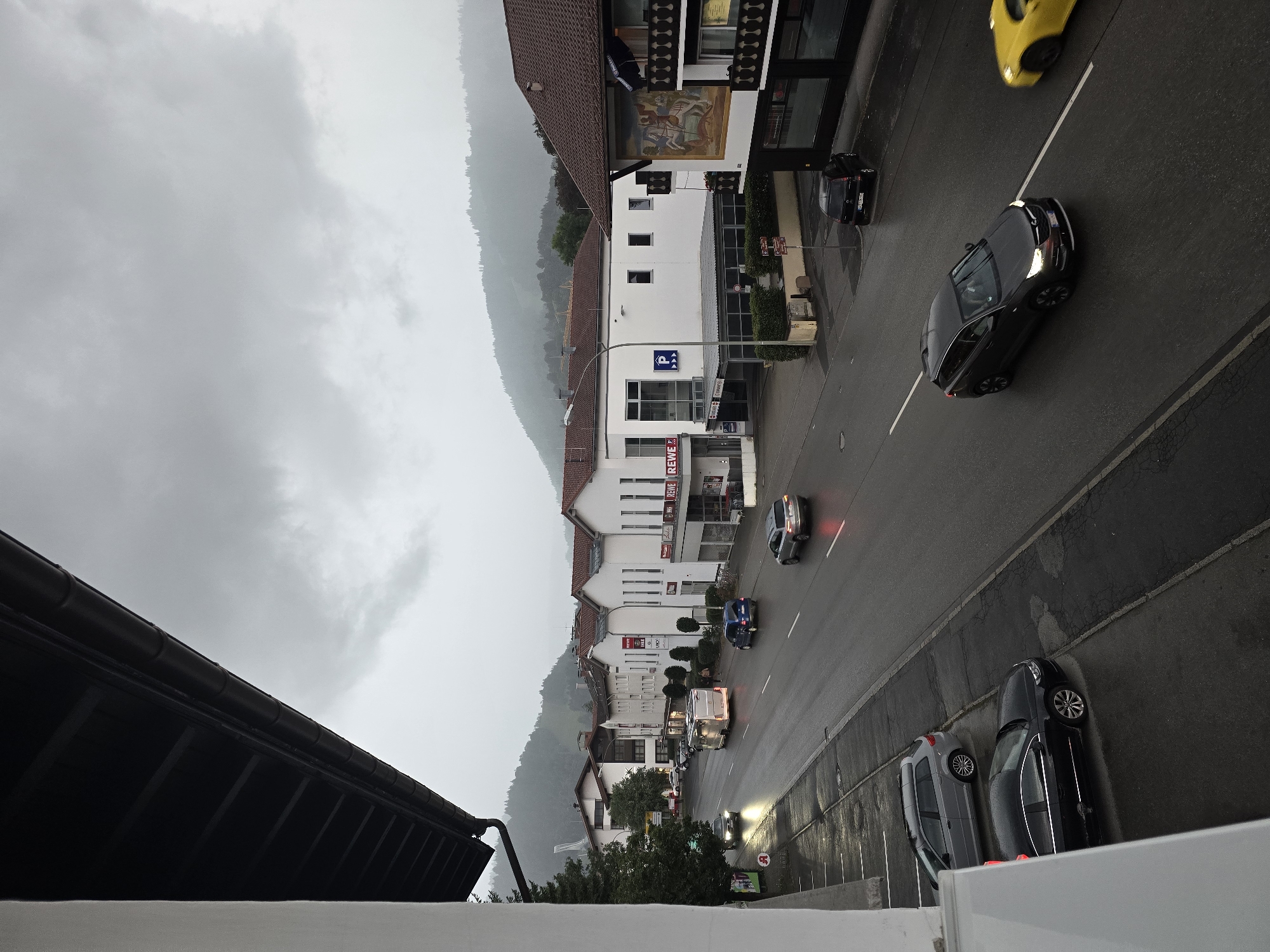 This is a vertical, wide-angle, outdoor shot of a street scene in Garmisch-Partenkirchen, Germany, on a cloudy day. The perspective is from a balcony overlooking the street. A white wall is on the left, and a dark-brown gutter system is visible overhead. The street is wet, indicating recent rain.
In the foreground, a black sedan is parked on the left side of the street, with its headlights on. To its right, a light gray station wagon is also parked. The street itself has two lanes of traffic. A dark gray SUV is driving in the right lane towards the viewer. To its right, a yellow sports car is also driving in the right lane, blurred due to motion.
Further down the street, a dark blue minivan is driving in the left lane. A white motorhome is in the right lane, with its brake lights illuminated. Another white sedan is driving in the left lane. A light-colored sedan is parked on the right side of the street, further down.
Several buildings line the right side of the street. The closest building has a dark brown roof and features a large mural depicting a knight on horseback. The building to its left is white with a dark brown roof and several windows. A sign with a blue "P" indicates parking. Another white building with a dark brown roof and multiple windows is visible further down. A sign for "REWE" is visible on one of these buildings.
The background shows a green, forested hill rising behind the buildings, with clouds obscuring the top of the hill. A street light is visible on the right side of the street. The street has white lane markings and a pedestrian sidewalk on the right.