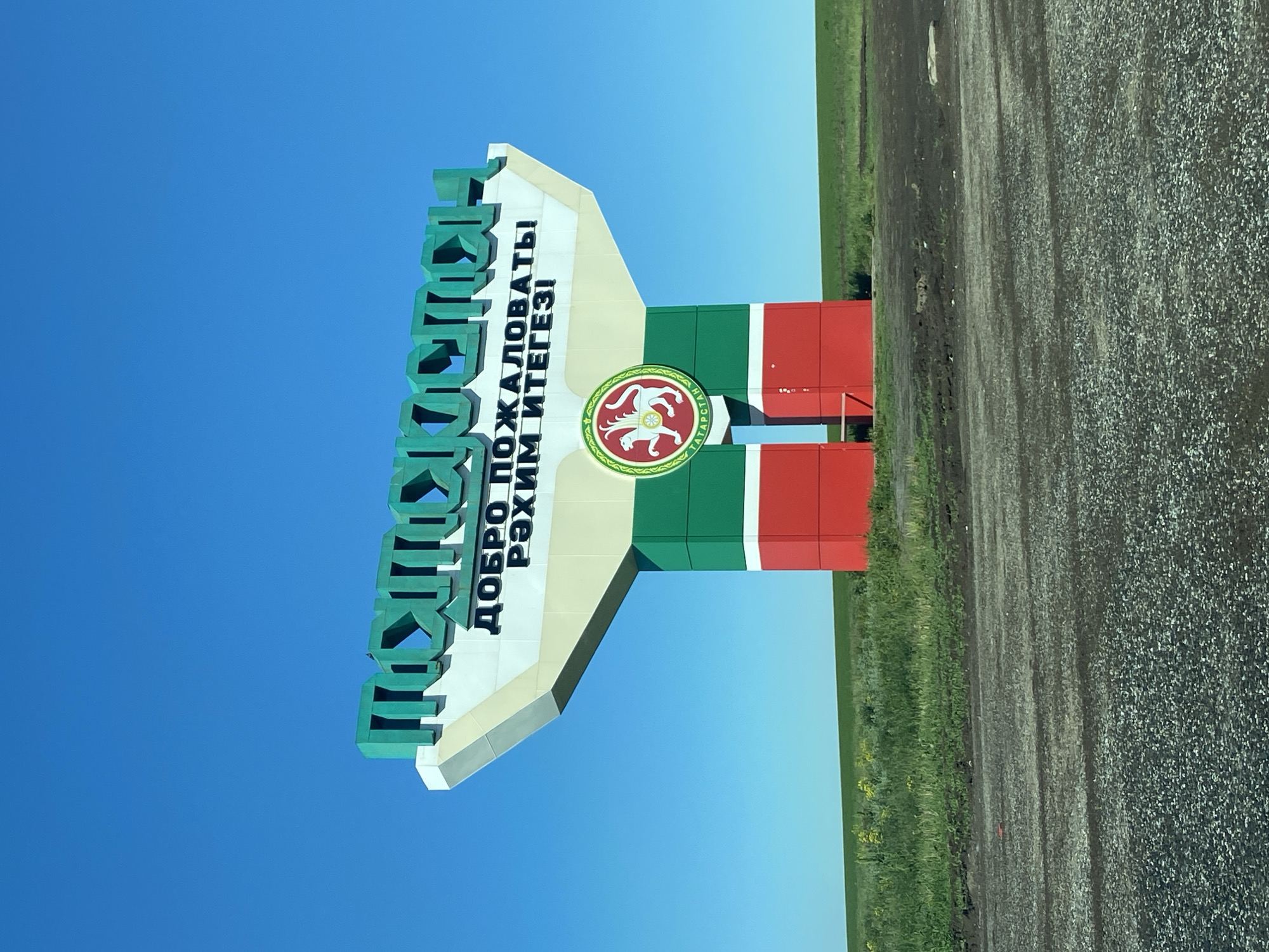 The image shows a sign for the city of Kamaevo, Russia, set against a clear blue sky. The sign is green on top, with the city's name in Russian and Tatar languages. Below the name, there is a section in beige, containing the phrase "ДОБРО ПОЖАЛОВАТЬ! РӘХИМ ИТЕГЕЗІ" (Welcome in Russian and Tatar). Below that, the sign is painted in the colors of the flag of Tatarstan: green, white, and red. On the white stripe, the coat of arms of Tatarstan is featured. The sign is situated next to a road, with gravel and grassy areas visible to the right.