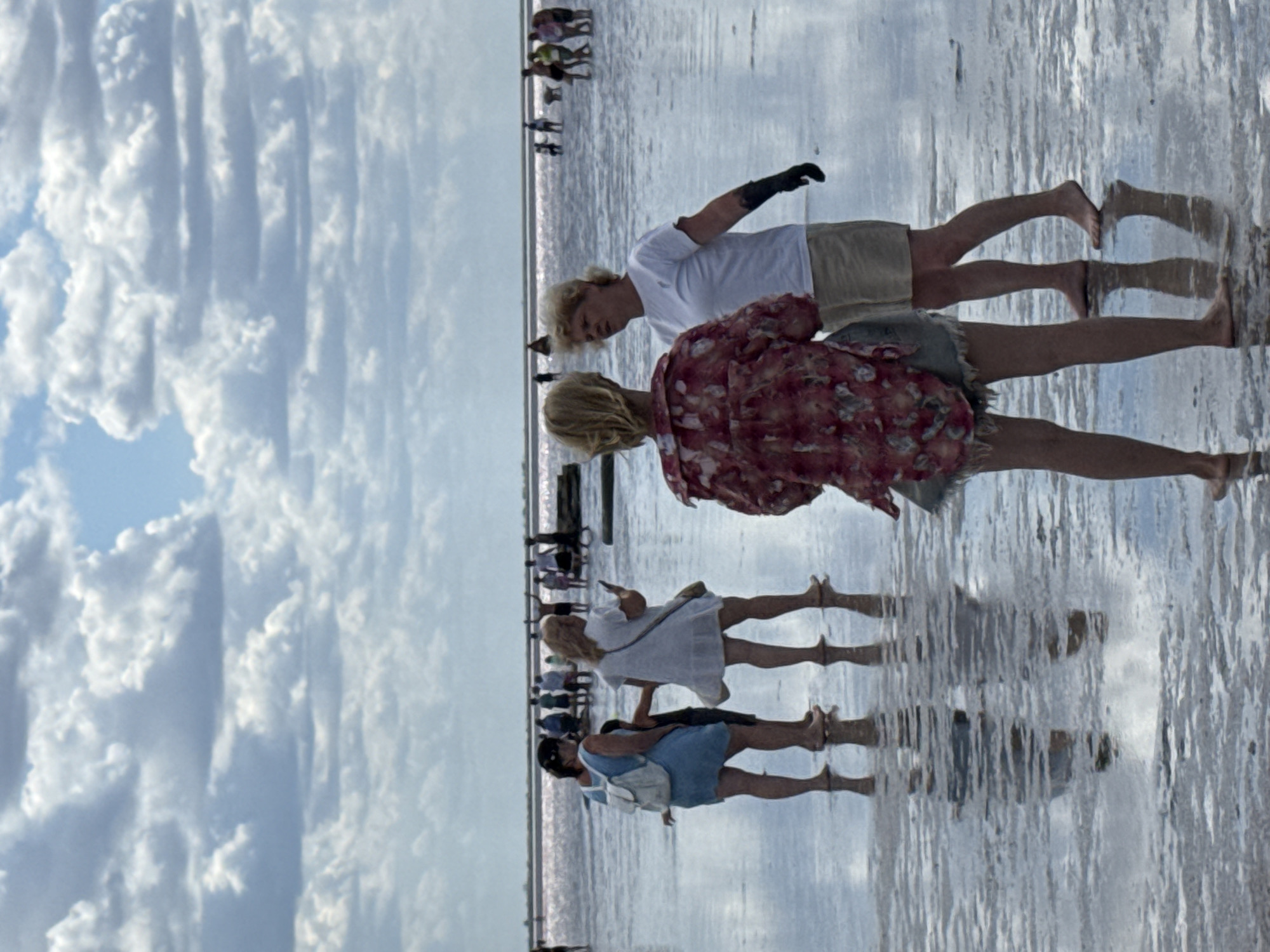 In Бурсоль, Russia, a group of people are gathered at a shallow, reflective body of water, likely a salt lake, given the pinkish hue of the distant horizon. The sky above is filled with dramatic, puffy white clouds against a pale blue background. In the foreground, four individuals are prominent. Two women, one in a blue dress with a white backpack and another in a white dress with a crossbody bag, stand facing away from the camera, with a child between them. To their right, two other women are engaged in conversation. One, wearing a floral print jacket and denim shorts, faces away from the camera. The other, in a white t-shirt and beige shorts, turns slightly towards the first woman, wearing a black glove on her right hand. The water's surface is highly reflective, mirroring the sky and the figures. In the background, numerous other people are spread across the water, some standing, others walking. The scene depicts a leisurely day in a unique natural environment.