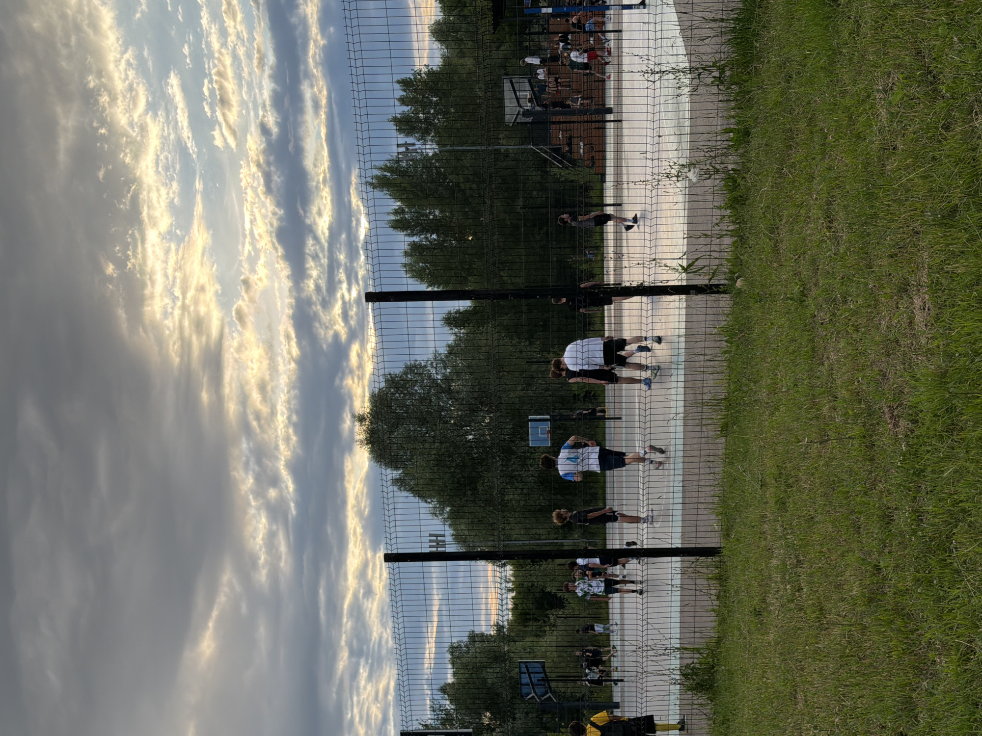 This is a vertical, slightly low-angle shot of a fenced outdoor sports court. In the foreground, a chain-link fence runs diagonally across the frame, with lush green grass to the right and the court to the left. The court appears to be asphalt or a similar hard surface with painted lines.
Several people are visible on the court, engaged in what seems to be a casual sports activity, possibly basketball or a similar game, given the presence of a blue basketball hoop in the background. Some individuals are standing, while others are in motion, suggesting an ongoing game or practice. They are dressed in athletic attire.
The background features a dense line of trees, indicating a park or recreational area. Above the trees, the sky is filled with dramatic, wispy clouds, illuminated by the warm, golden light of either sunrise or sunset. This suggests the time of day is either early morning or late evening. The light on the clouds creates a striking contrast with the darker trees and the shaded areas of the court.
The overall impression is one of an active outdoor scene set against a beautiful, dynamic sky. There is no visible text in the image. The location context of Kazan, Russia, is provided but not explicitly discernible from the visual elements themselves.