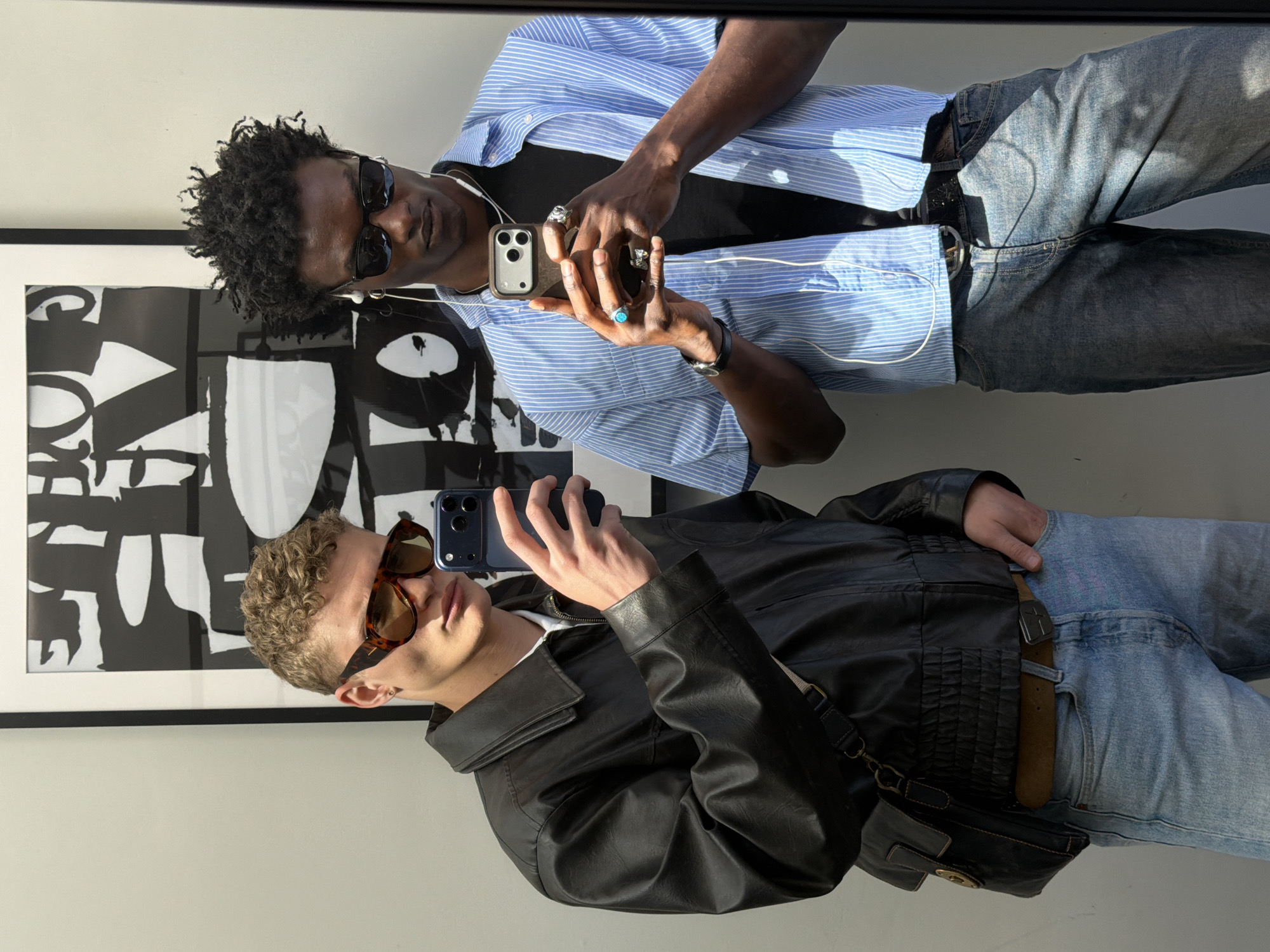 The image depicts two young men taking mirror selfies in an indoor setting in London, United Kingdom. The background features a white wall and a large framed abstract art print in black and white.
On the left, a man with dark skin and short, curly black hair wears black sunglasses, a blue and white striped short-sleeved shirt over a black t-shirt, and light blue jeans. White wired earphones are visible around his neck. His hands are adorned with several rings, including one with a turquoise stone, and a black wristband. He holds a phone with a light-colored case, pointed towards his reflection.
On the right, a man with light skin and curly blonde hair wears tortoiseshell sunglasses and a black leather jacket over a white top. He is dressed in light blue jeans secured with a brown belt. A black shoulder bag with an attached smaller black pouch hangs across his body. He also holds a phone, which has a dark case, facing the mirror.
Both individuals have neutral, focused expressions as they interact with their phones. The bright lighting suggests it is daytime. The abstract art print displays bold, non-representational black shapes and brushstrokes on a white background, without forming any clearly legible text.