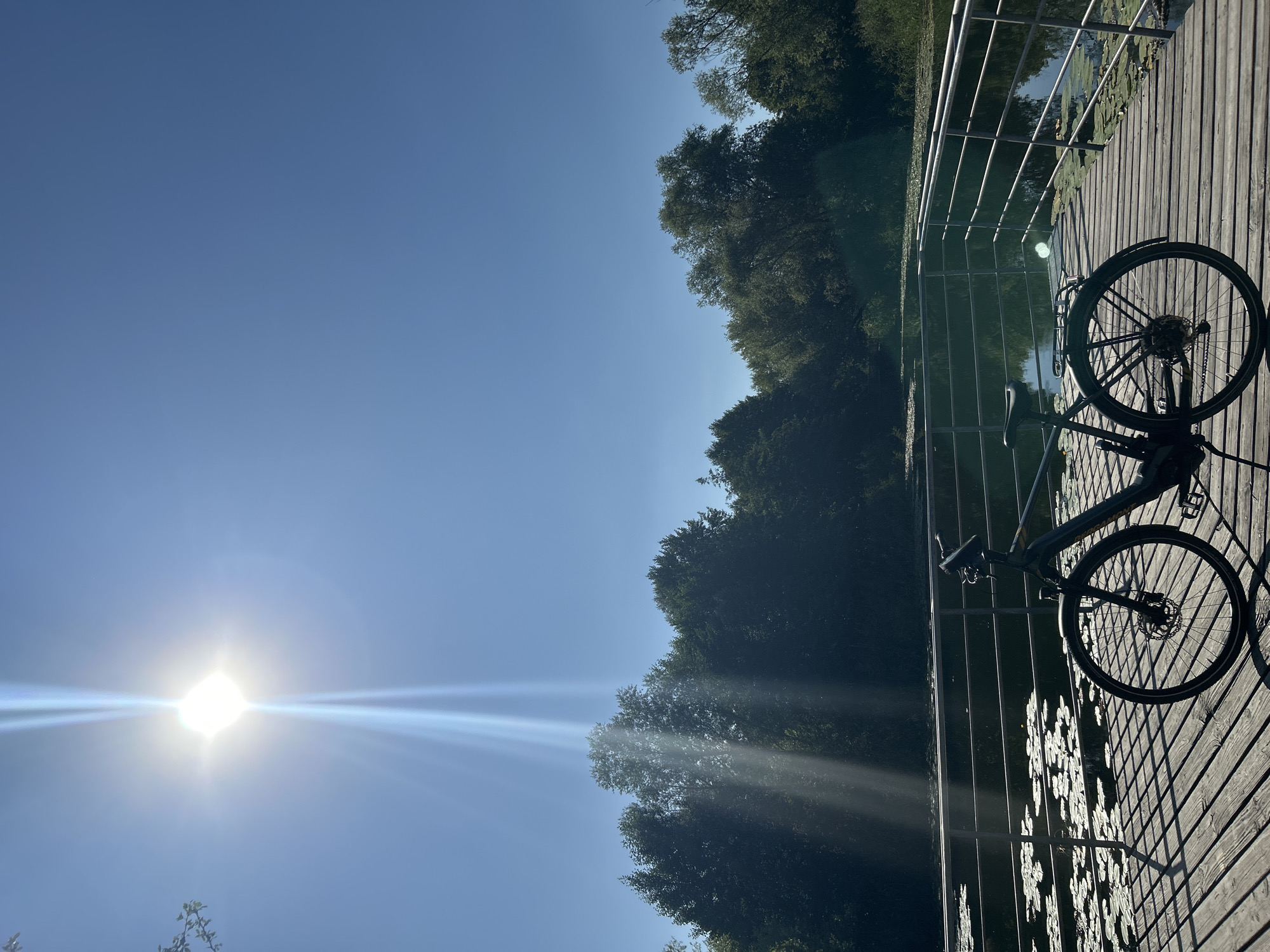 An electric bicycle is parked on a wooden pier next to a body of water. The sun is shining brightly in a clear blue sky, casting lens flares and bright beams of light downwards. The water is covered in lily pads, and dense trees line the far bank. A metal railing runs along the edge of the pier.