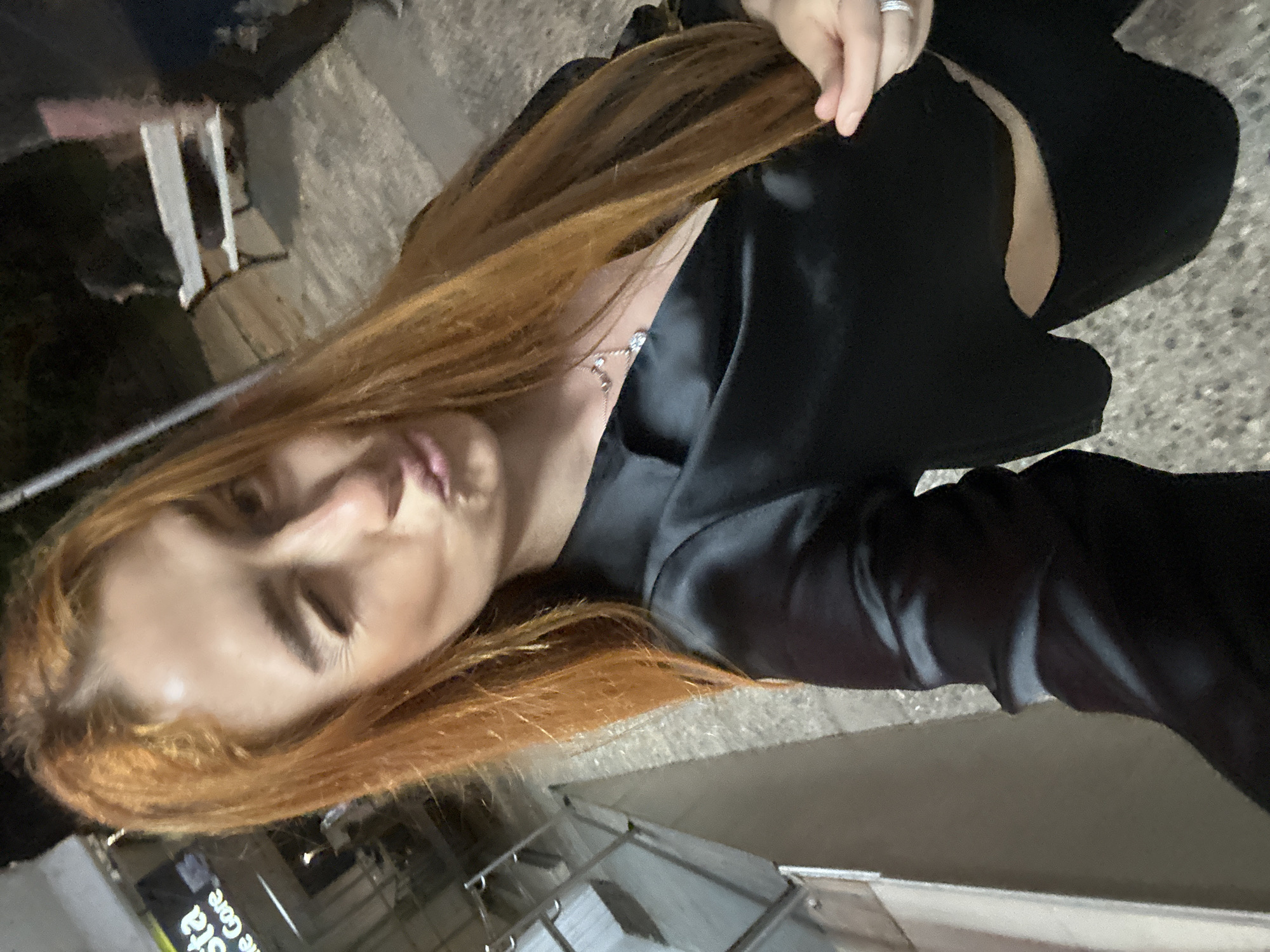 The image captures a selfie of a young woman at night in an urban setting. She is seen from a high angle, with her head tilted and her long, reddish-brown hair flowing downwards. Her right hand, adorned with a silver-colored ring, is lightly touching her hair. She is wearing a dark, possibly black, satin or silky top, which appears to feature a design with a cut-out on the arm, revealing skin. A delicate, silver-toned necklace with small, sparkling elements hangs around her neck. Her expression features a slight pout, with her eyes looking downwards, suggesting she is gazing at the camera (likely a phone held by herself).
The background is dark and out of focus, indicating the photo was taken at night or in low light conditions. Visible background elements include blurred concrete structures, indistinct metal railings, and what appears to be part of a building or street. In the lower left, a blurry sign with white text on a dark background is partially legible, displaying "sta" and "the core". The overall impression is a casual, personal moment captured in an urban environment in Budva, Montenegro.