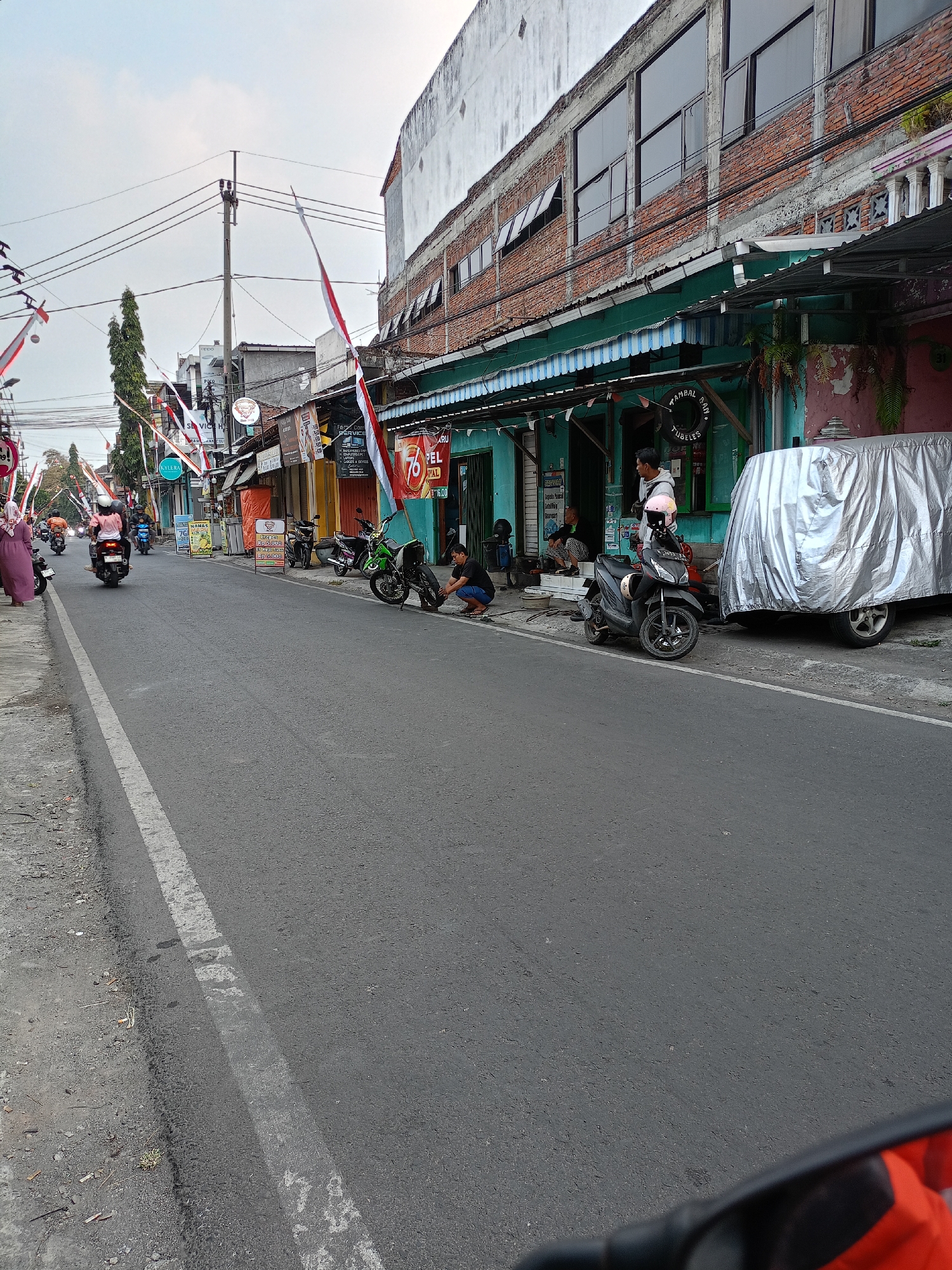 The image captures a street scene in Batu, Indonesia, likely during a festive occasion, as evidenced by the numerous Indonesian flags (red and white) decorating the street. The scene unfolds on a paved road with a white line marking the lane. On the right side of the road, a row of buildings with turquoise lower facades and exposed brick upper sections line the street. Some of these buildings appear to be shops or businesses, with awnings extended and signs visible. One building has a large silver tarp covering a vehicle parked in front of it.
Several motorcycles are present: a green one parked near the foreground, a black scooter with a pink helmet on the seat, and other motorcycles further down the street, some with riders. People are interacting in the scene. In the foreground on the right, a person wearing a dark shirt and blue shorts is kneeling and appears to be working on the green motorcycle's rear wheel. Further back, a group of people are gathered near the entrance of one of the buildings. On the left side of the street, a person in a purple hijab is walking away from the camera, and other people on motorcycles are seen riding away. The sky is overcast, suggesting it might be early morning or late afternoon. The environment appears to be an urban street with pedestrian and vehicle traffic.