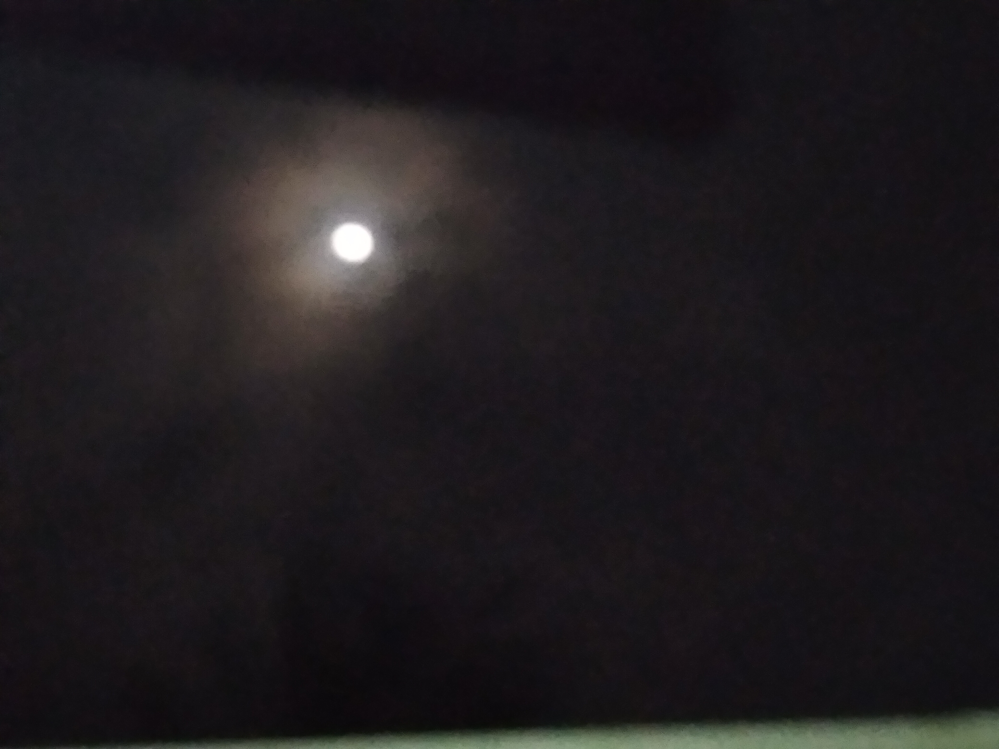 A bright, full moon is visible in a dark, cloudy sky. The moon is slightly off-center to the right and appears luminous. The clouds are diffuse and create a hazy effect around the moon. The bottom left portion of the image shows a blurred green object, possibly a piece of vegetation or a building element. The rest of the image is dark, indicating it was taken at night. The scene is likely outdoors.