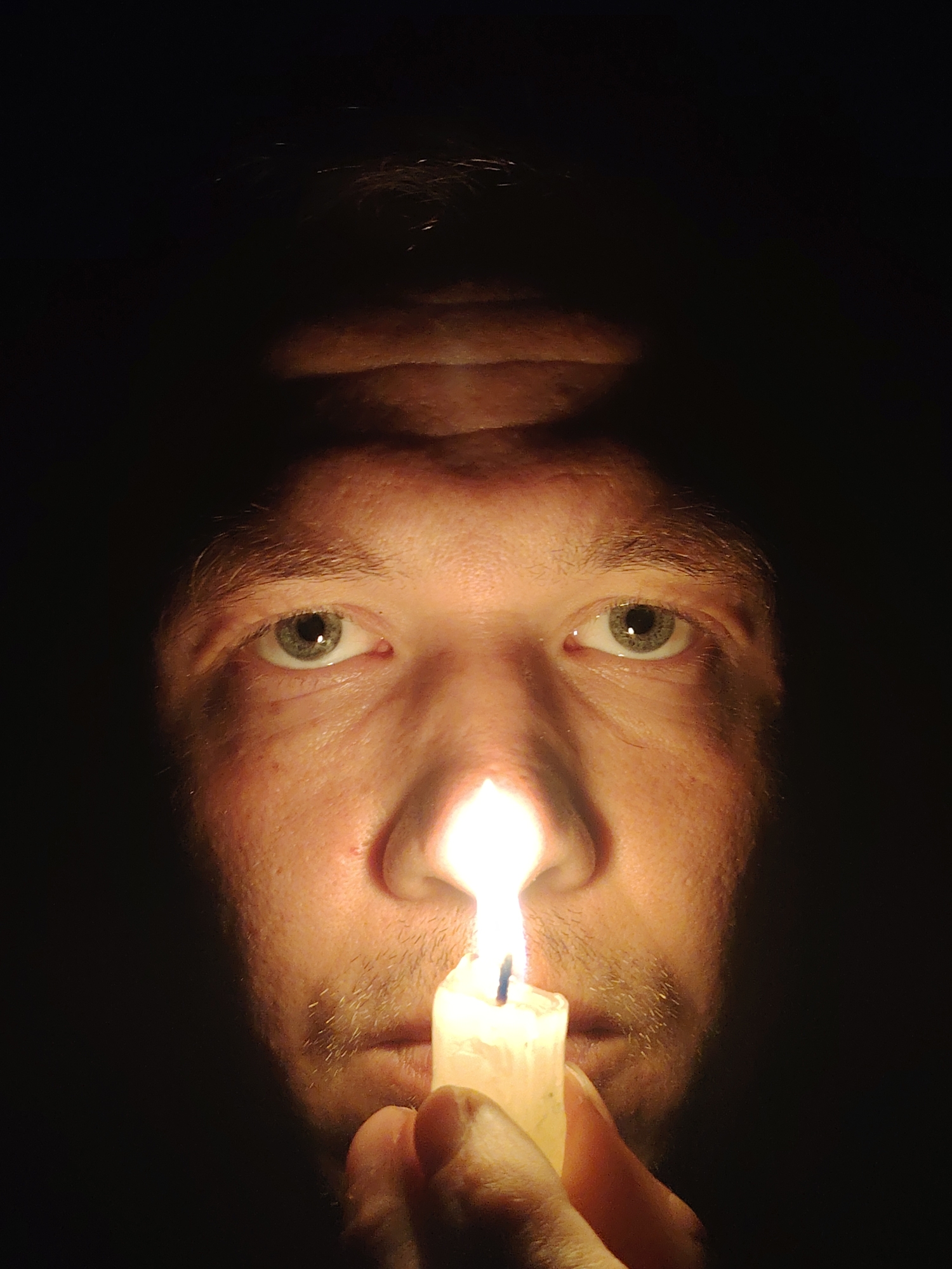 This is a close-up, low-angle shot of a person's face illuminated by a candle held in their hand. The background is entirely dark, suggesting a nighttime or indoor setting with minimal ambient light. The subject's eyes are open and looking directly at the camera, and their forehead shows some horizontal wrinkles. A thin mustache and stubble are visible on their upper lip and chin. The candle itself is partially visible, held in the subject's right hand, with its flame creating a bright light source that casts shadows and highlights on the face. The image appears to be a self-portrait. There is no information in the image to suggest a location in Chaikovsky, Russia.
