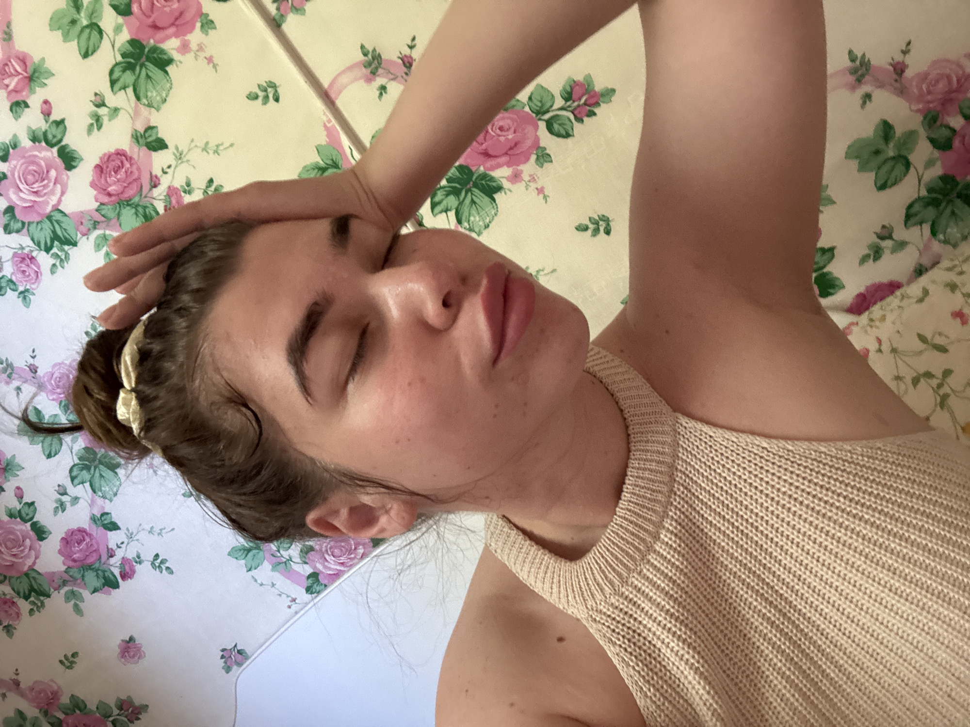 A young woman with her eyes closed leans her head on her hand, her arm bent at the elbow. She has her hair pulled up in a messy bun with a gold scrunchie. She is wearing a beige knitted top. The background is a floral wallpaper with pink roses and green leaves. The image appears to be taken indoors, possibly in a bedroom or living room. No specific location in Мещёры, Russia can be identified from the image.