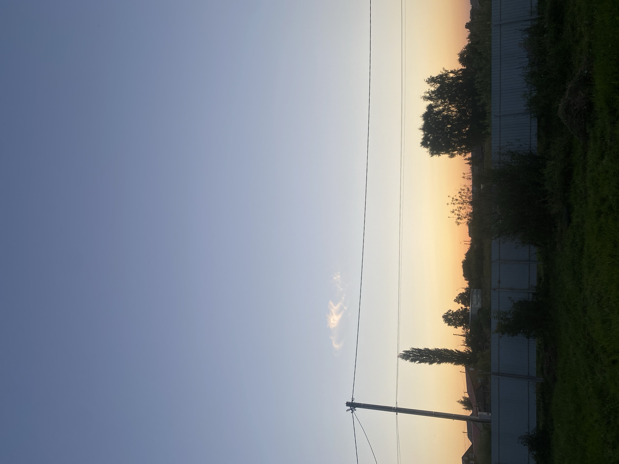 The image captures a twilight scene in the village of Tolbazy, Russia, with a clear sky transitioning from blue to a soft orange gradient near the horizon. A thin, wispy cloud is visible high in the sky. In the middle ground, a telephone pole stands to the left, with wires stretching horizontally across the frame. A tall, slender tree is positioned behind the pole, followed by a line of foliage and trees forming a silhouette against the colorful sky. A light-colored fence runs horizontally across the lower portion of the image, separating the foreground from the background. The foreground consists of dark green grass, with some uncut hay or dry grass in the lower right corner. Scattered houses and structures are visible behind the fence and trees on the left side of the image, suggesting a rural or semi-rural residential area. The overall atmosphere is serene and quiet, typical of a countryside evening.