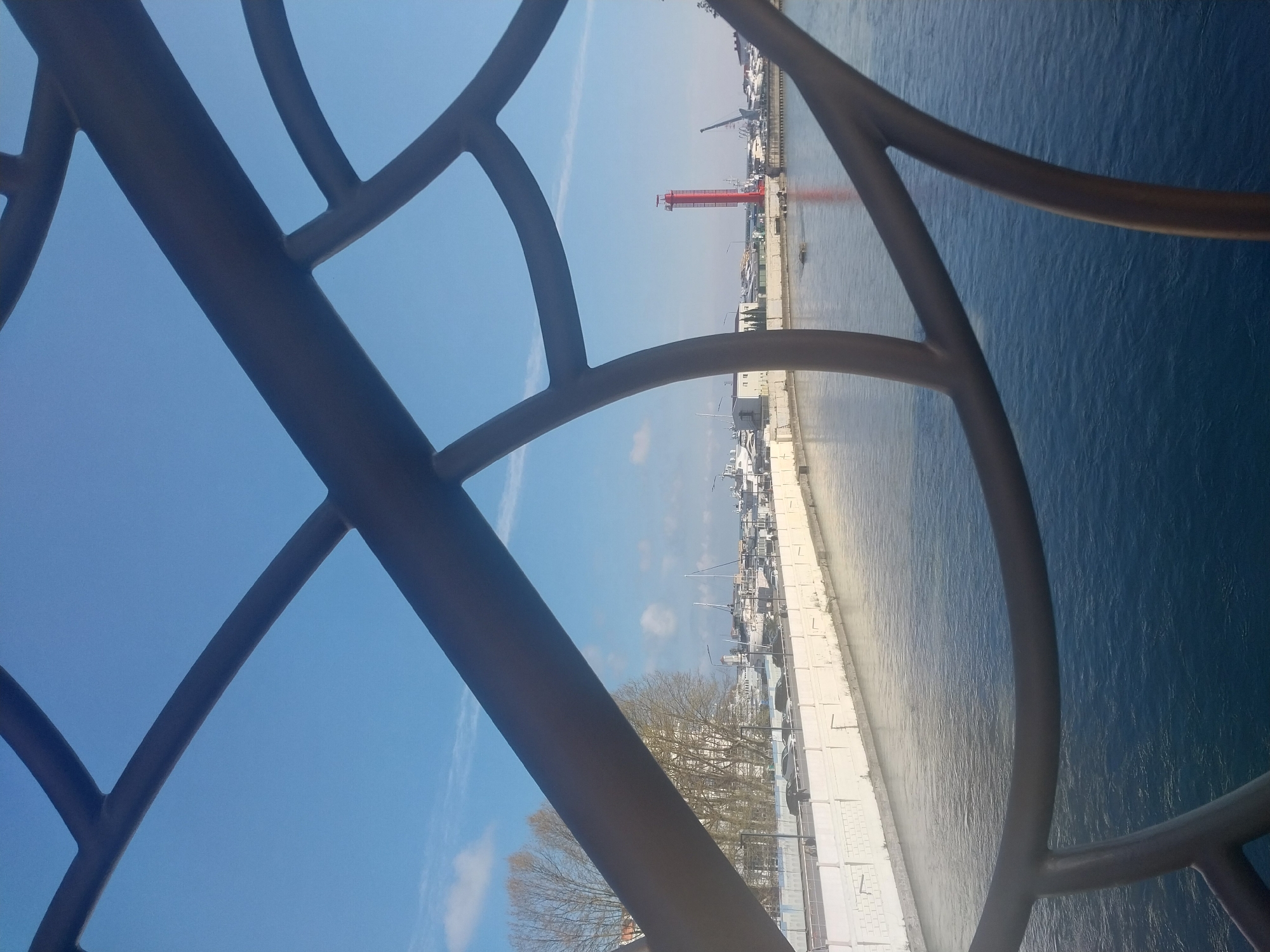 An elevated view captures a serene marina scene, framed by a large, dark grey, curved metal structure in the foreground, likely a railing or architectural element. The sky above is a clear, bright blue with a few faint, wispy clouds, indicating a sunny day.
To the right, a calm body of water, possibly the Black Sea, reflects the light with gentle ripples. A long, light-colored concrete or paved pier stretches along the water's edge, hosting numerous docked boats and yachts of various sizes.
A prominent tall, slender red structure, possibly a lighthouse or a distinctive harbor tower, stands in the mid-ground, its reflection clearly visible in the water. Beyond the pier, more waterfront buildings and structures can be seen. On the landward side, a cluster of bare-branched trees suggests a time of year outside of peak foliage.
The scene is devoid of visible human activity or text. It presents a peaceful, sunny daytime panorama of a harbor, consistent with the coastal city of Sochi, Russia.