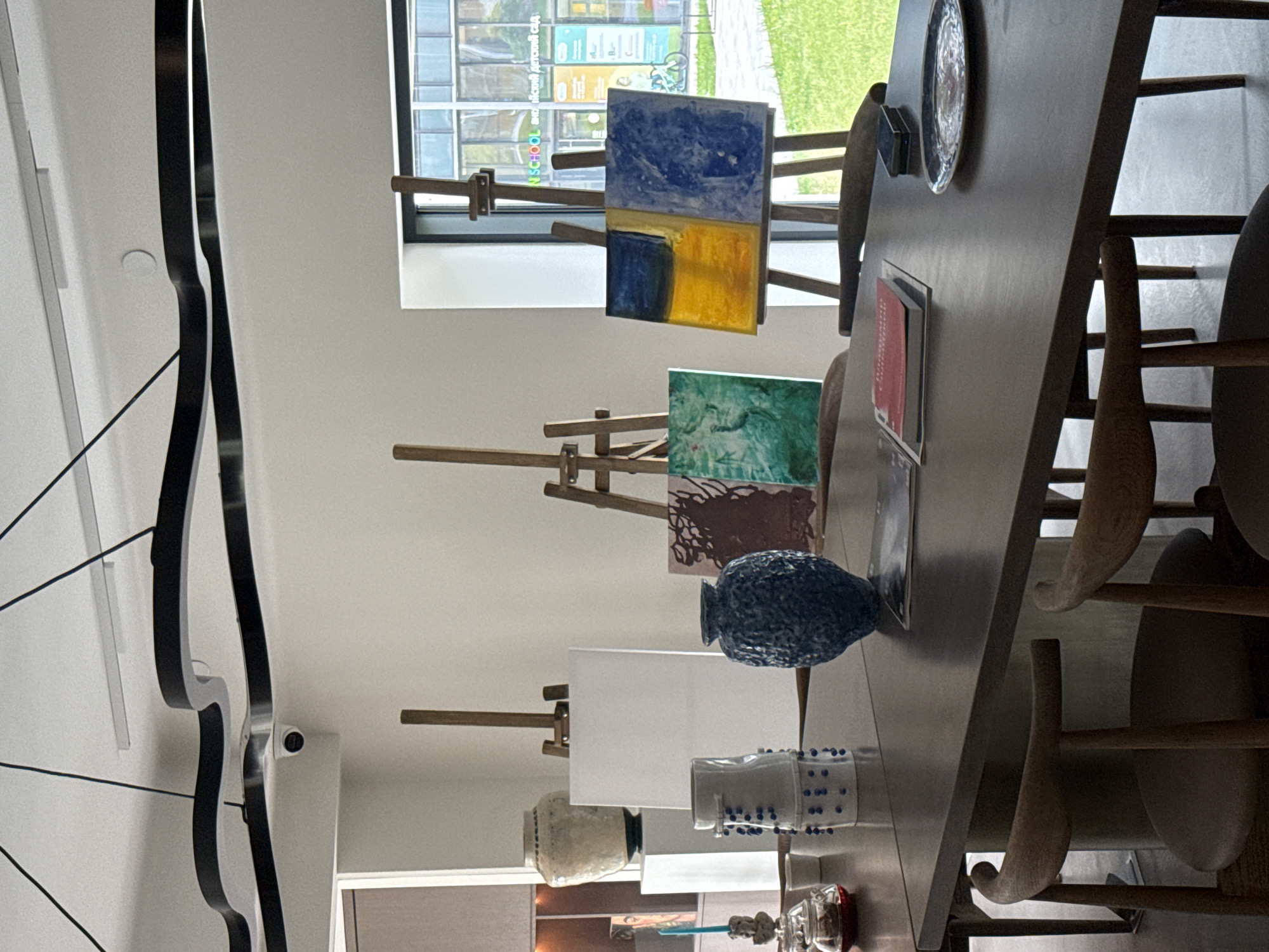 This image captures an indoor setting, likely a classroom or art studio, with a focus on a long, dark wooden table and various art supplies. The room is well-lit by natural light streaming through a large window on the far wall.
On the right side of the image, the long table is set with several wooden chairs featuring curved backs. Arranged on the table are three small wooden easels, each displaying a piece of art or a blank canvas. The easel closest to the window holds a small painting with distinct blue and yellow sections, reminiscent of the Ukrainian flag. Next to it, another easel displays a small, abstract painting primarily in green hues. The third easel, further down the table, holds a blank white canvas, suggesting ongoing or upcoming creative activity.
Further along the table, several decorative items are visible, including a dark blue, textured vase, a light-colored textured pot, and a clear container filled with blue beads or pins. A silver decorative plate and a small red book or folder are also on the table.
Through the window, a green lawn and a building can be seen outside. Crucially, reflections on the window glass reveal visible text: "SCHOOL" and "Детский сад" (Detskiy sad, meaning "kindergarten" or "preschool" in Russian), strongly indicating an educational environment in Moscow, Russia.
On the left side of the room, a modern, abstract black metal light fixture hangs from the white ceiling, complementing the contemporary aesthetic. A small white security camera is mounted on the wall near the ceiling. The overall scene suggests a creative learning space, possibly set up for an art class or workshop during daytime hours.