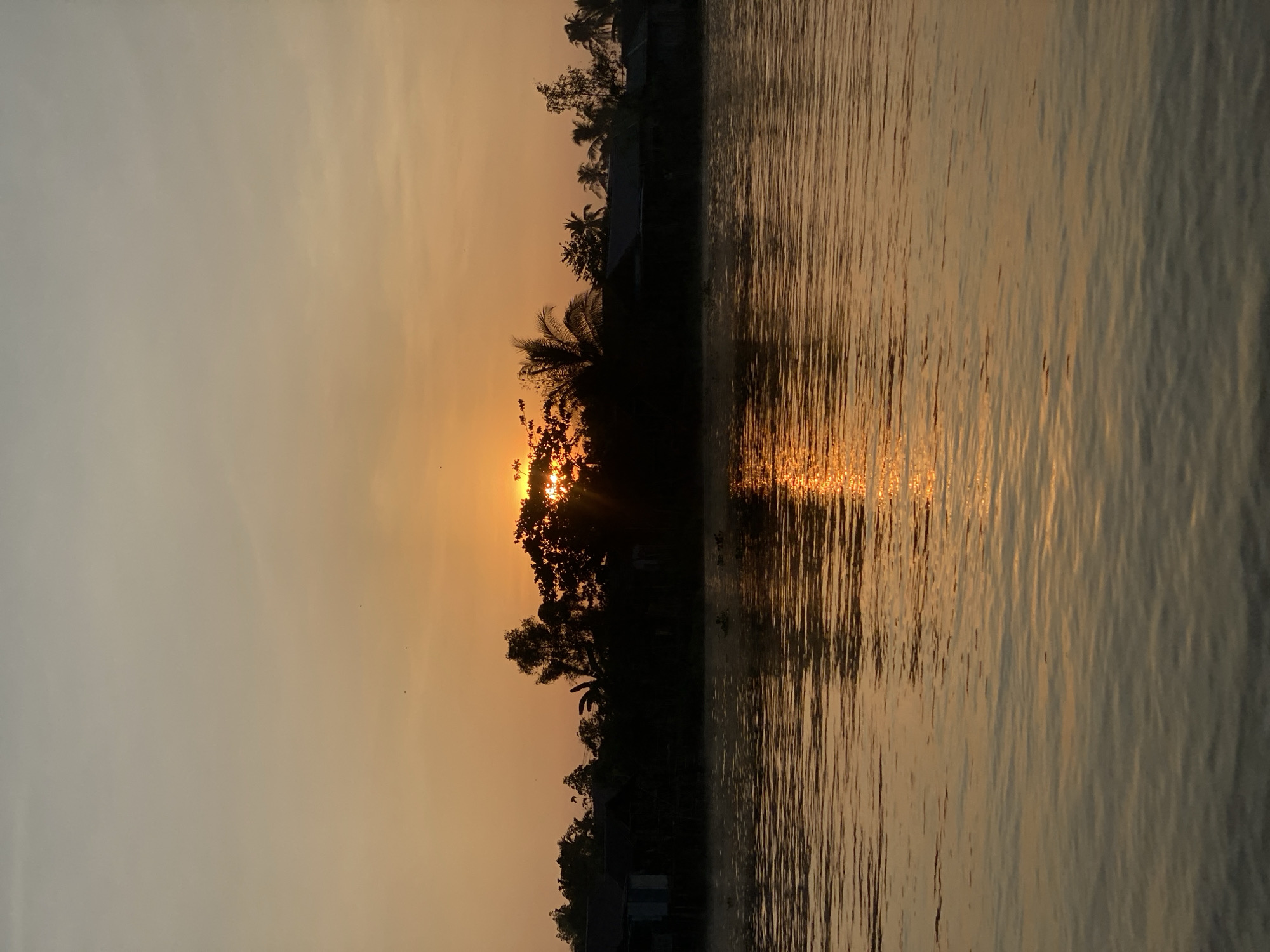 The image depicts a serene natural landscape at sunset. The sun, partially obscured by foliage, casts a warm, orange glow across the sky and reflects vividly on the calm waters of a river. Silhouetted against the sunset are trees and what appear to be small structures, possibly houses, lining the riverbank. The overall scene suggests a tranquil rural setting, likely in a location like Sungai Tandipah, Indonesia, known for its rivers and natural beauty. There are no people or explicit actions visible in the image; the focus is on the natural environment and the atmospheric quality of the sunset. The water is gently rippled, further enhancing the peaceful ambiance.